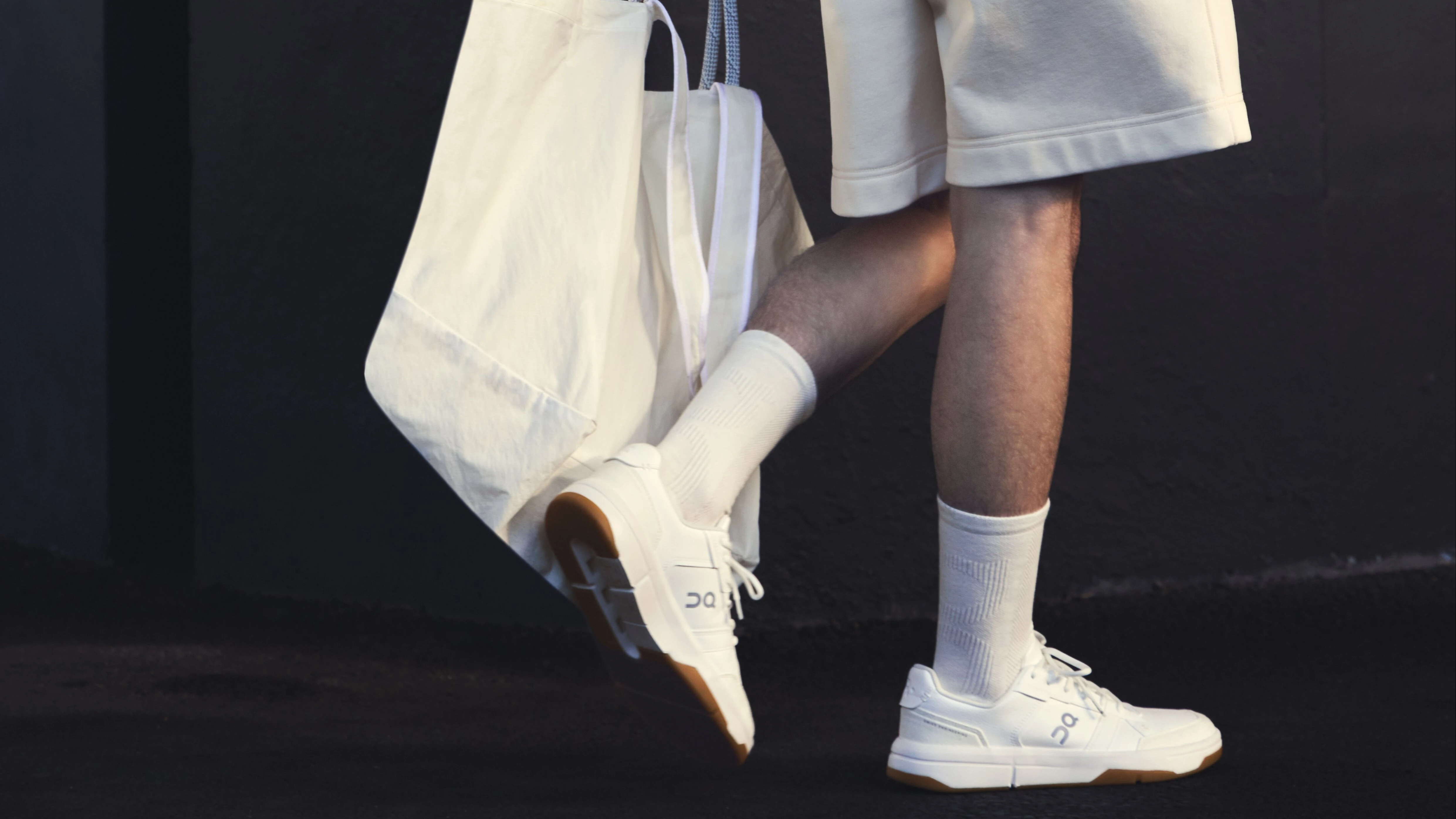 Eine Person von der Hüfte abwärts: Sie trägt Shorts, Socken und On Sneaker für den Alltag und in ihrer Hand eine Tote-Bag.