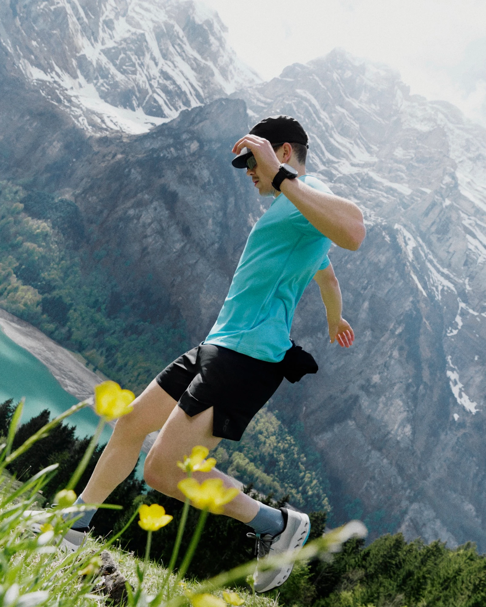 A runner running down a mountainside, mountains in the background and yellow flowers in the foreground.