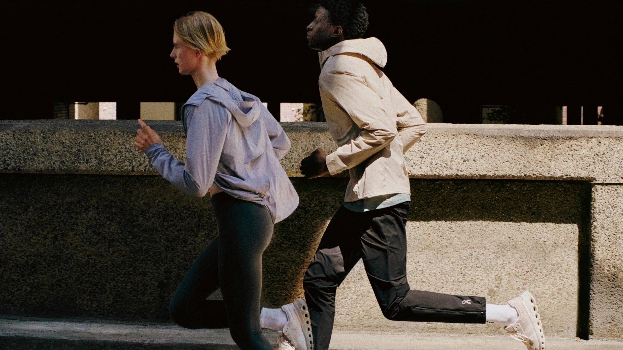 A woman runs slightly ahead of a man on a city sidewalk, both wearing On gear.