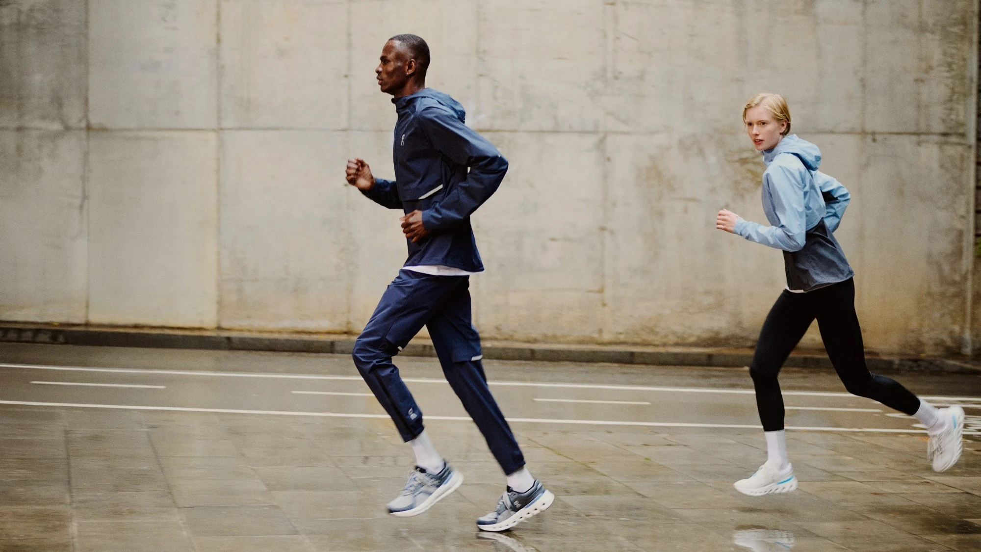 Two runners wearing On outerwear running on a wet street.