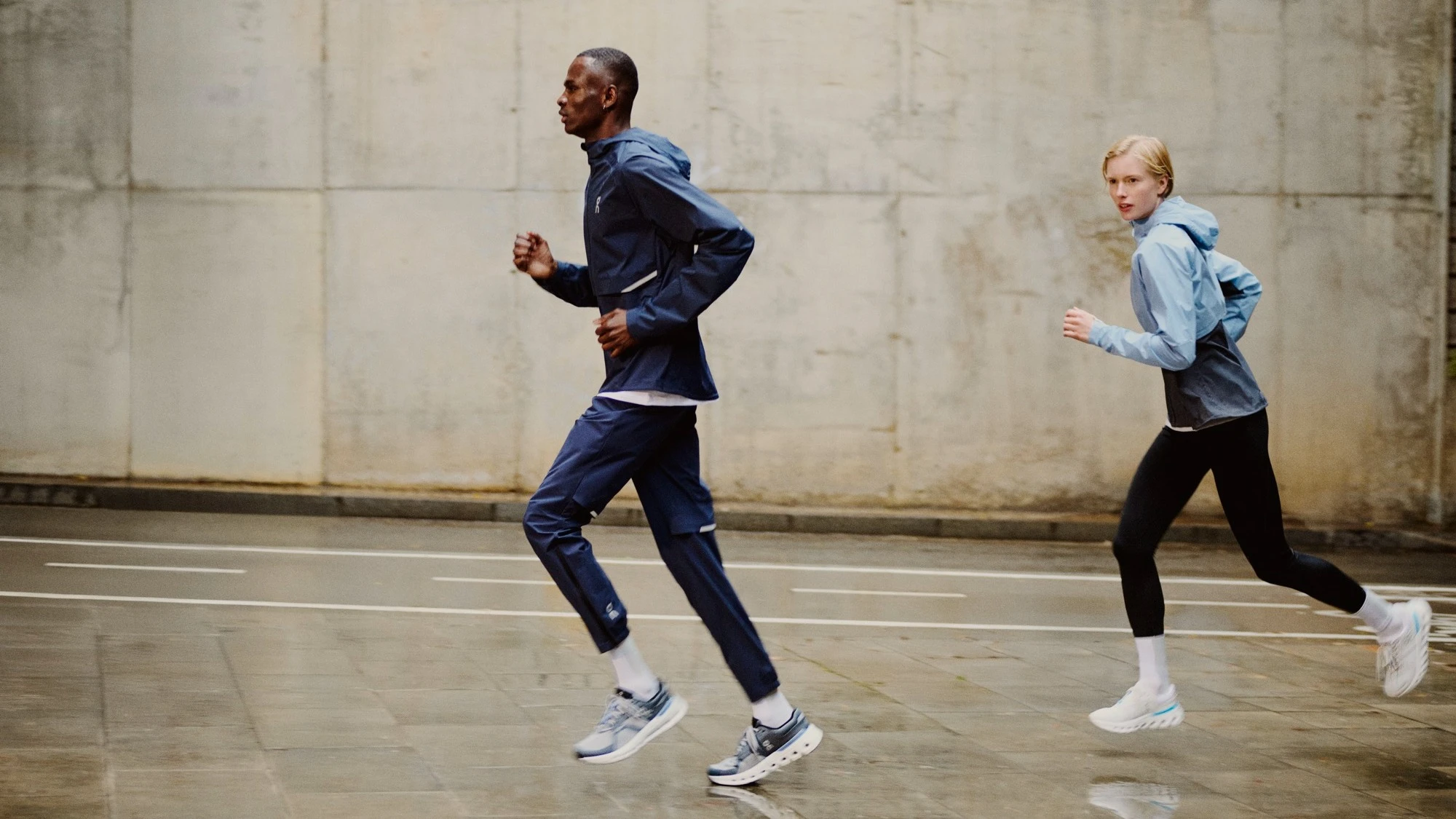 Two runners wearing On outerwear running on a wet street.