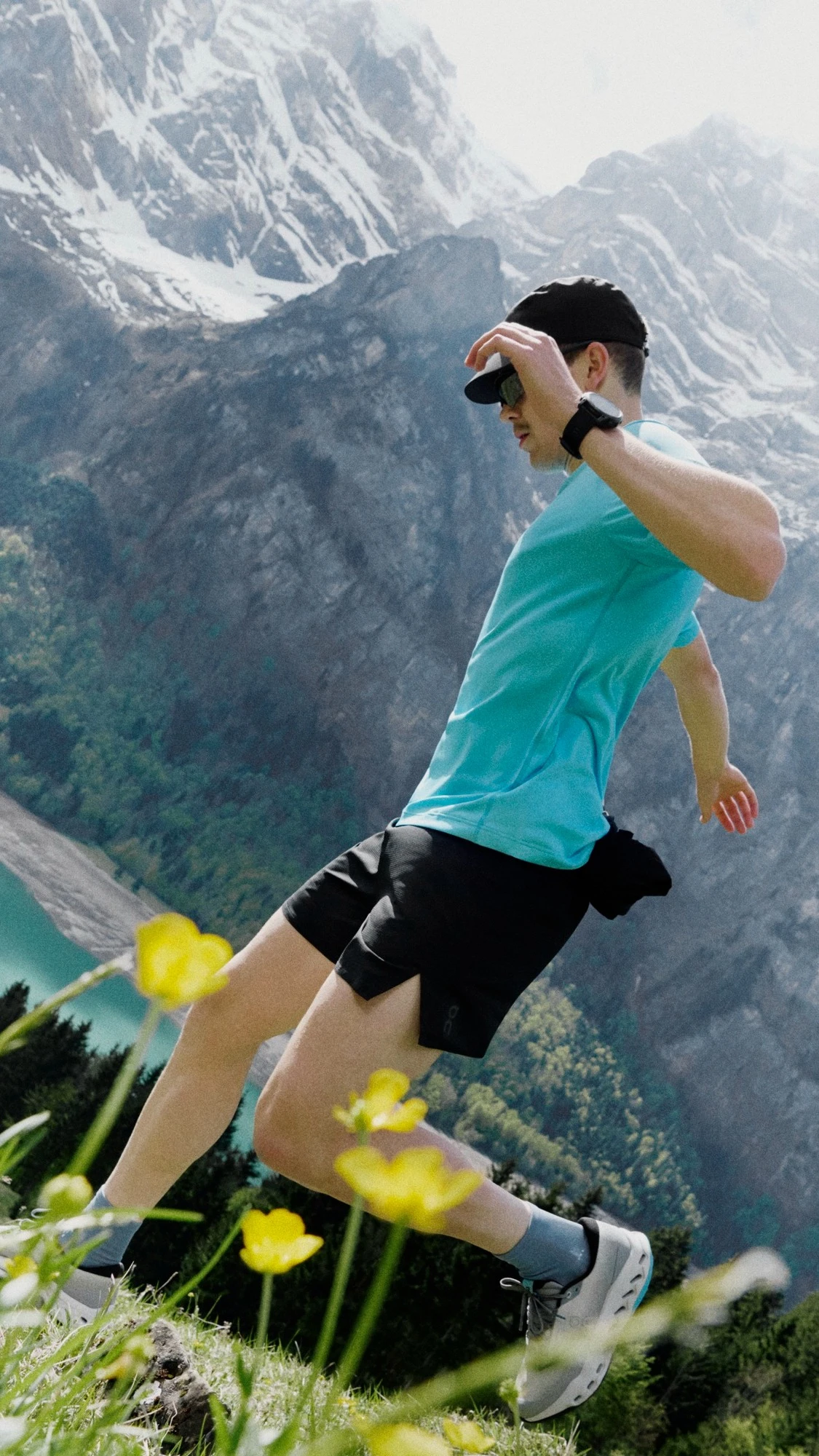 A runner running down a mountainside, mountains in the background and yellow flowers in the foreground.