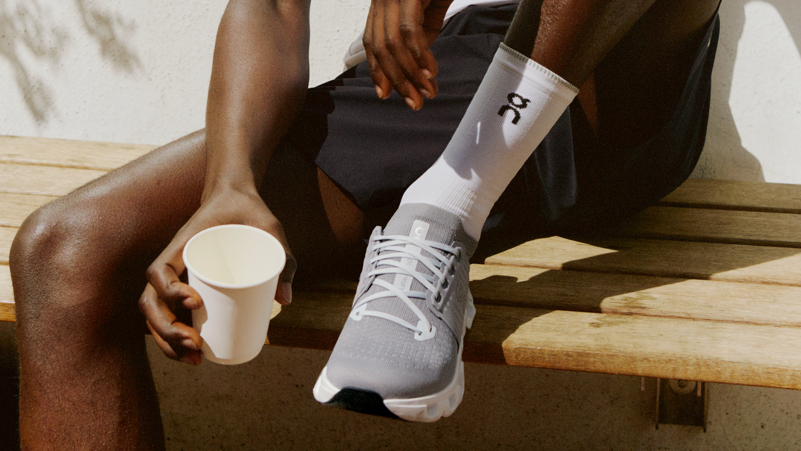 A person holding a cup of coffee while wearing On brand athletic shoes and workout clothes, preparing for a morning workout.