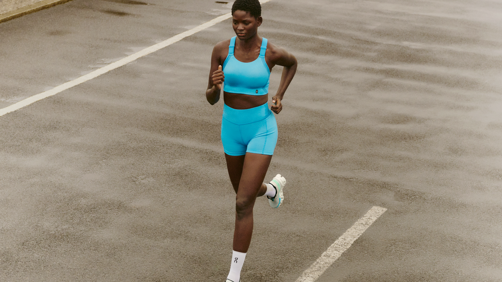 Una mujer de aspecto atlético corre en una pista con un sujetador deportivo y pantalones cortos azul claro. 