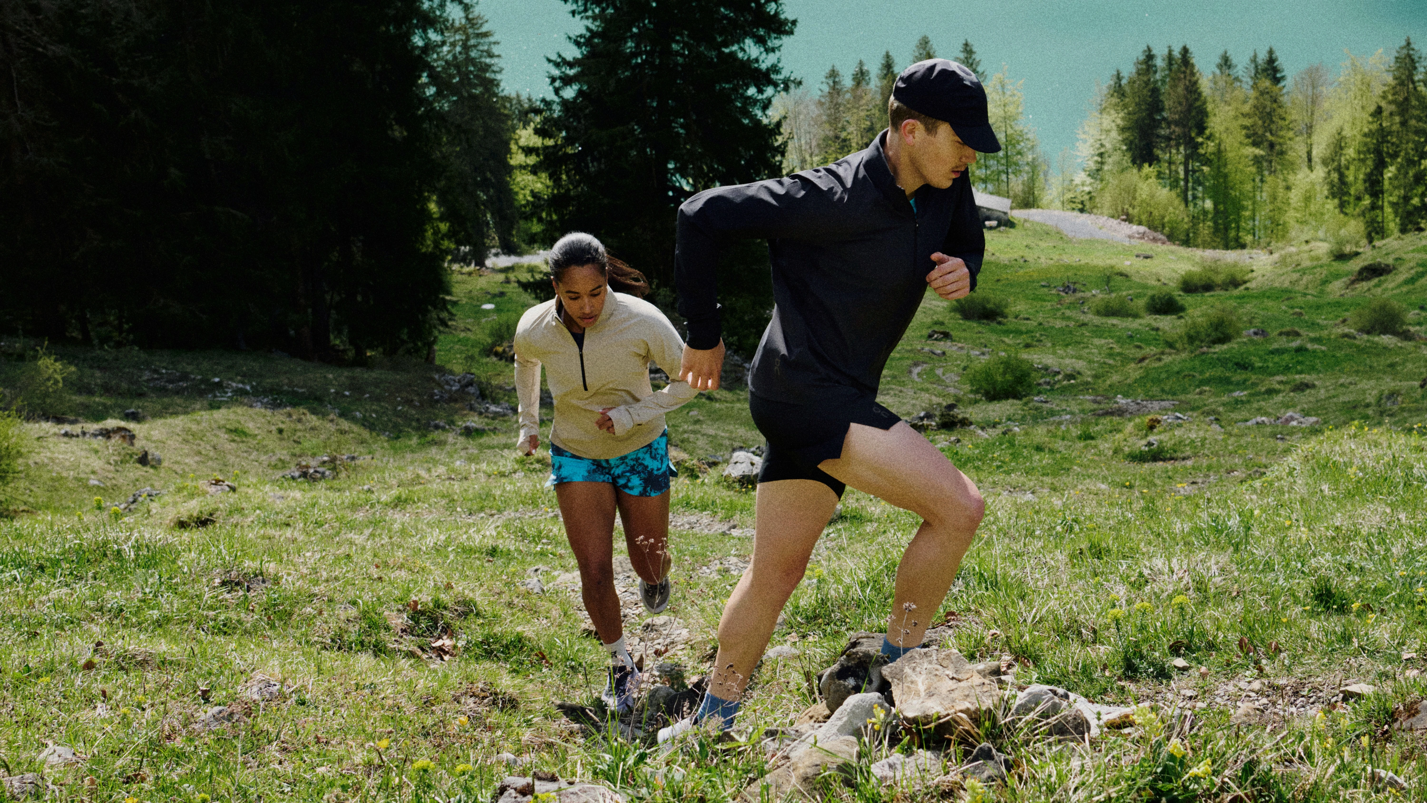 Two runners in On gear climbing a grassy mountain trail surrounded by trees.