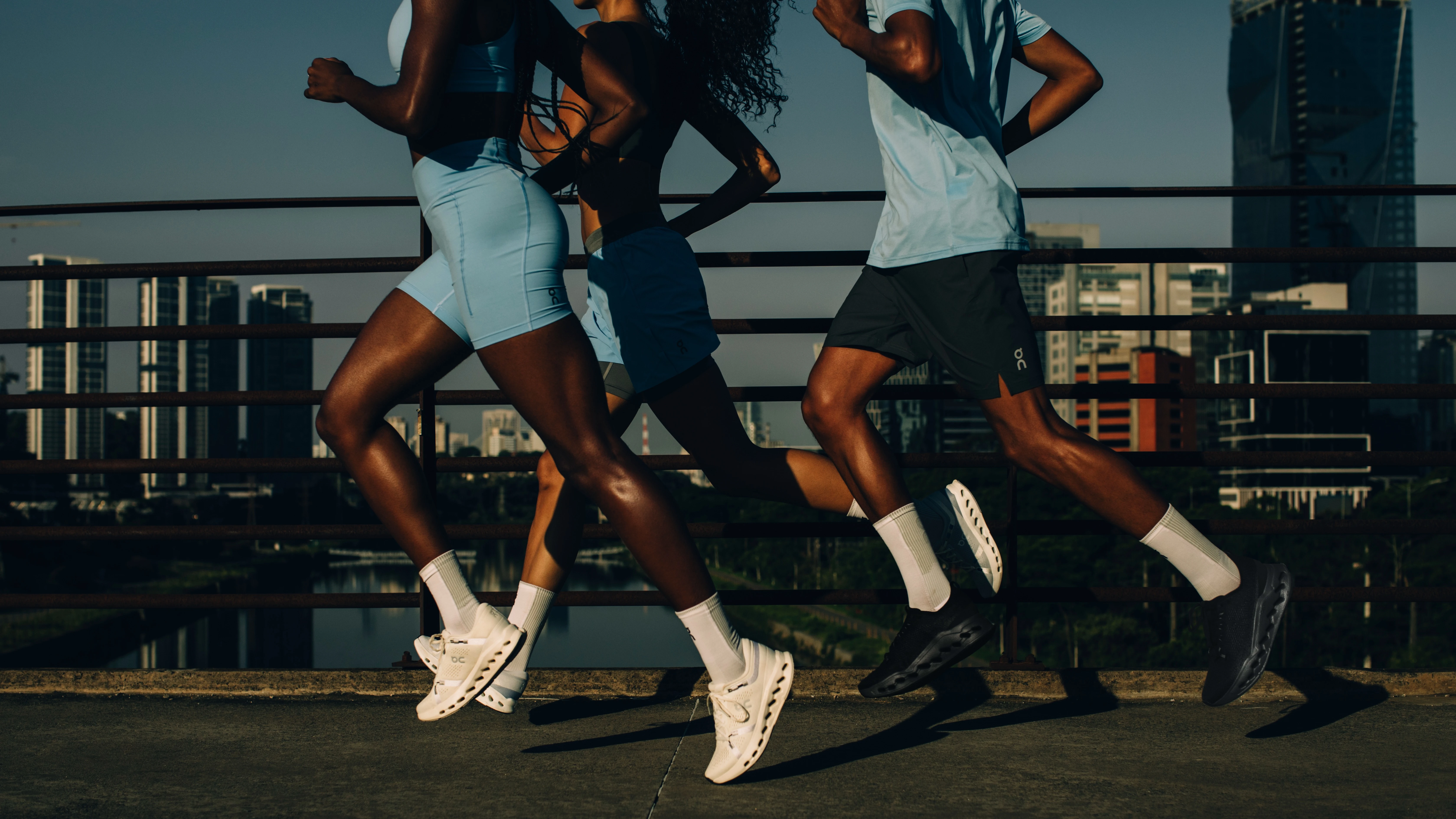 Three runners in On gear with their heads out of frame, running across a bridge in the city. 