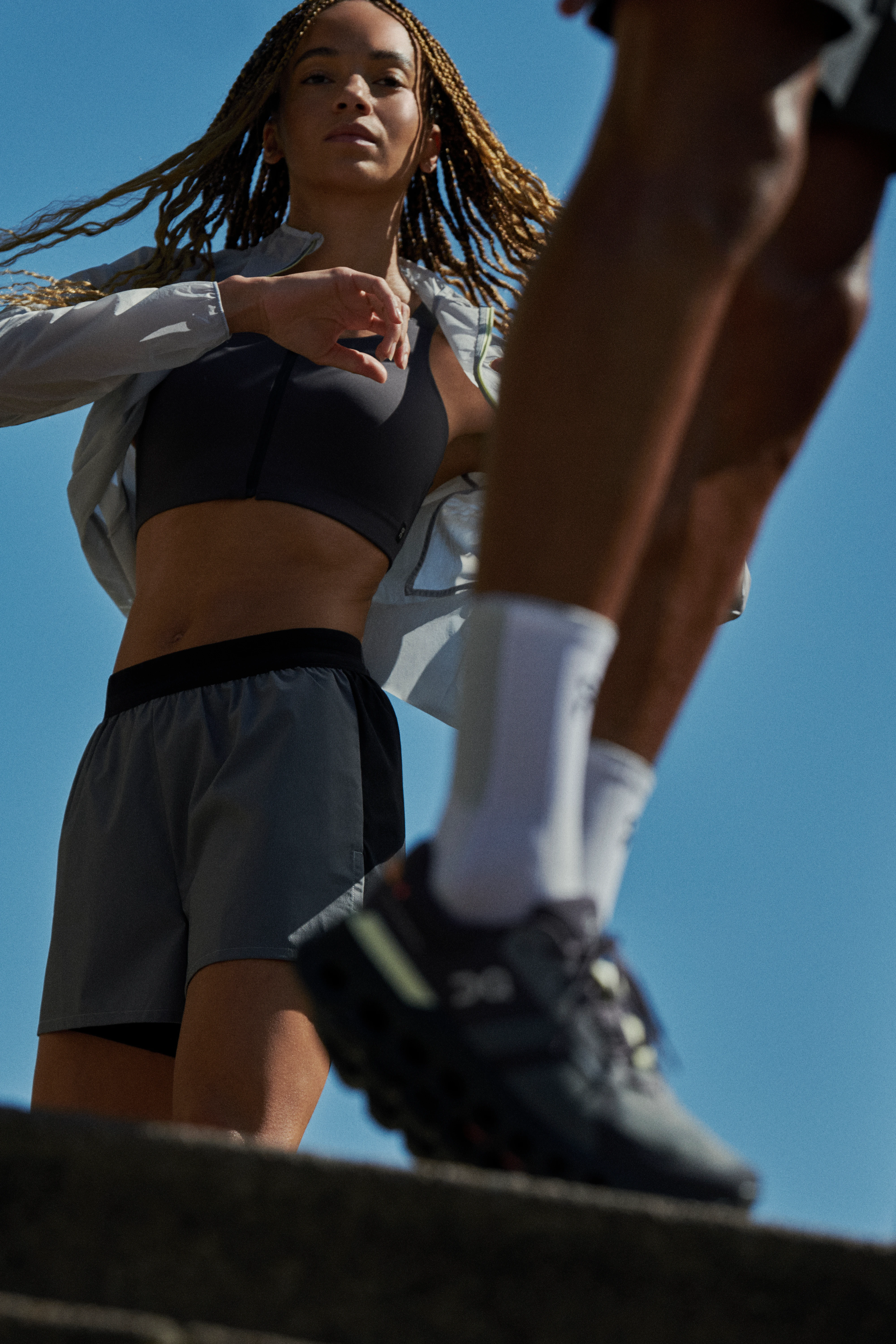A runner twists stretches in the background and a focus on calves and On running shoes in the foreground.