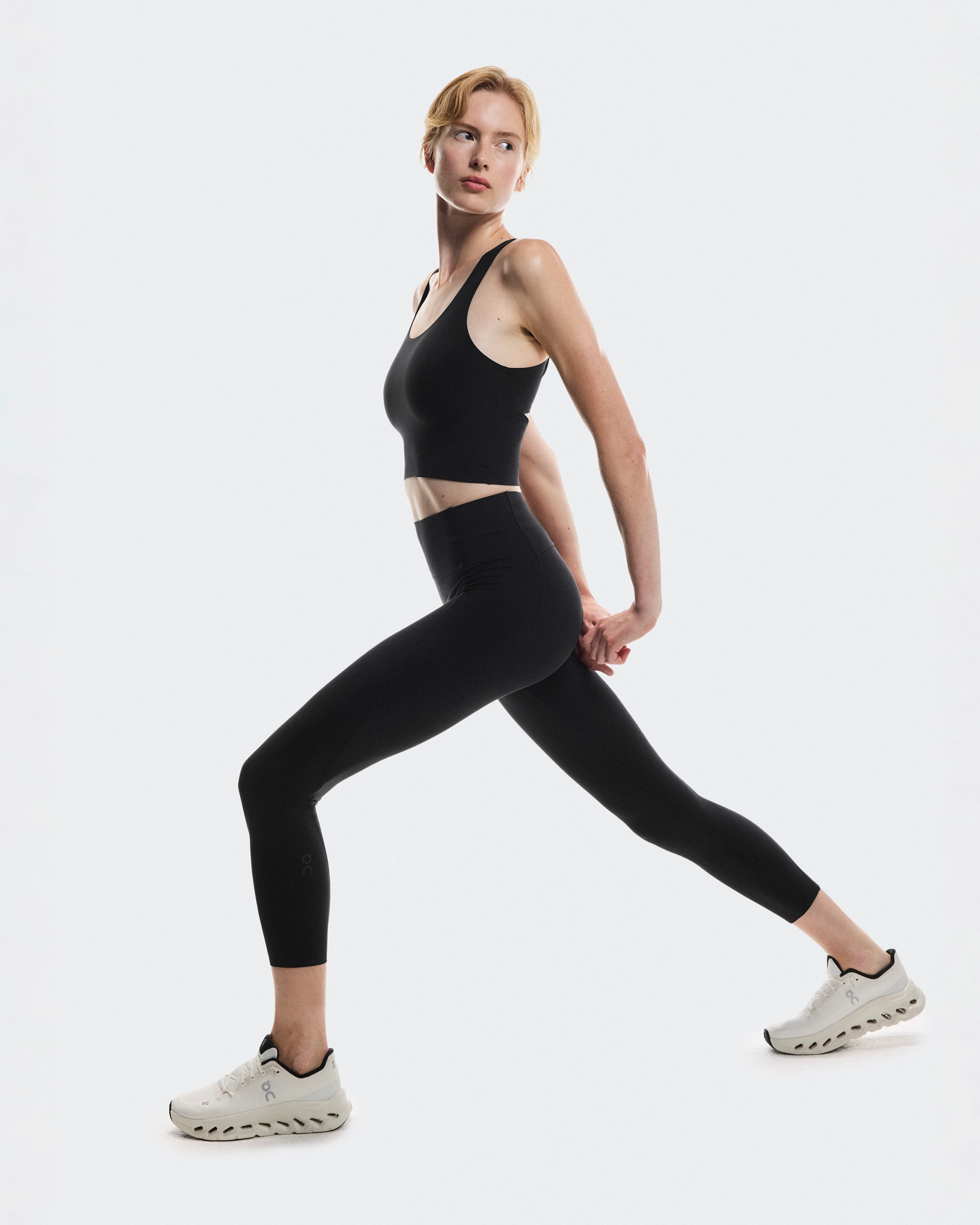 Woman in black cropped tank and matching leggings from On Running, stretching in a lunge position.
