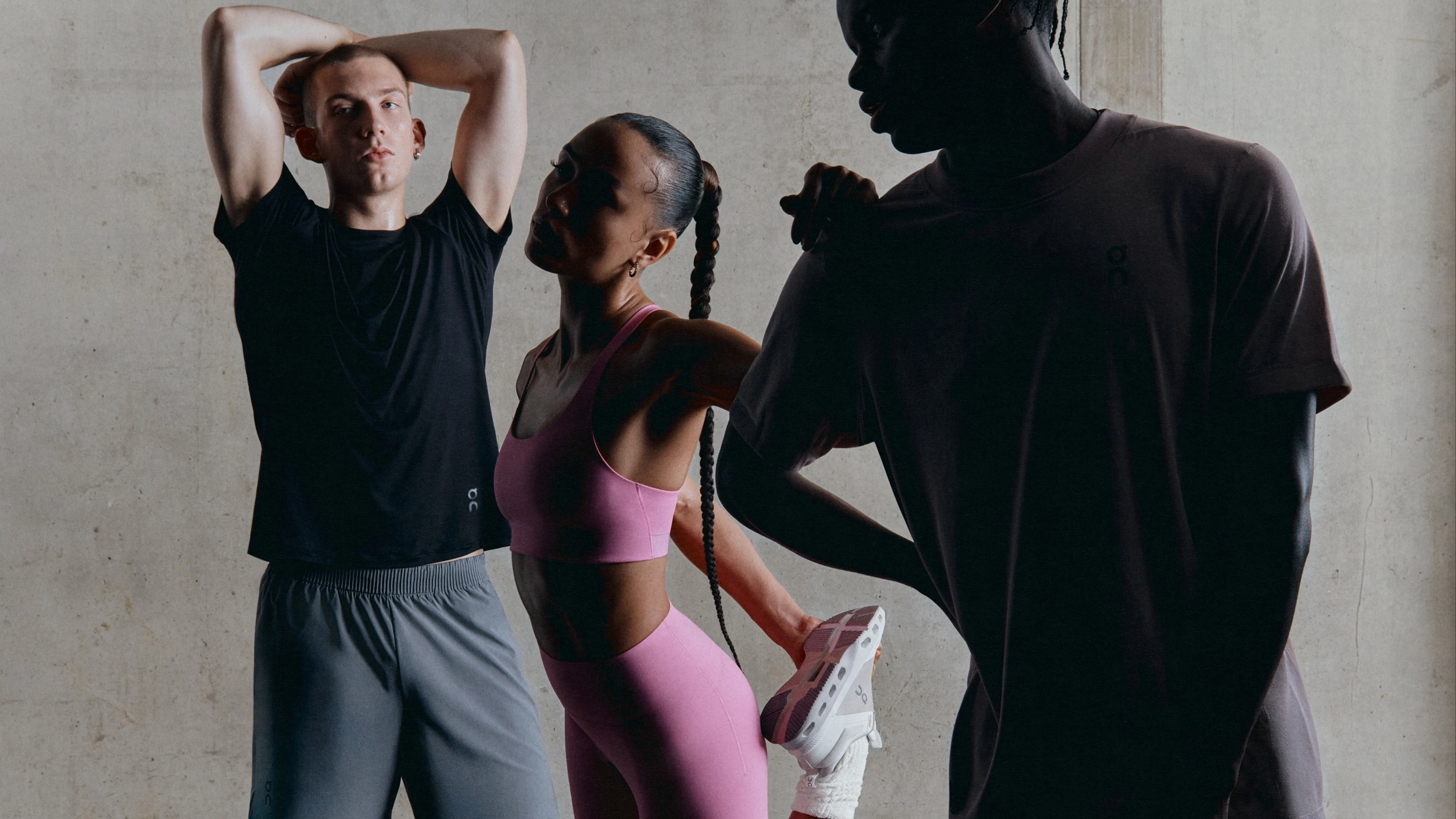 Three athletes in athletic wear stretch before performing physical activity. Two men are wearing neutral shorts and t-shirts, and the woman is wearing a pink sports bra and leggings.