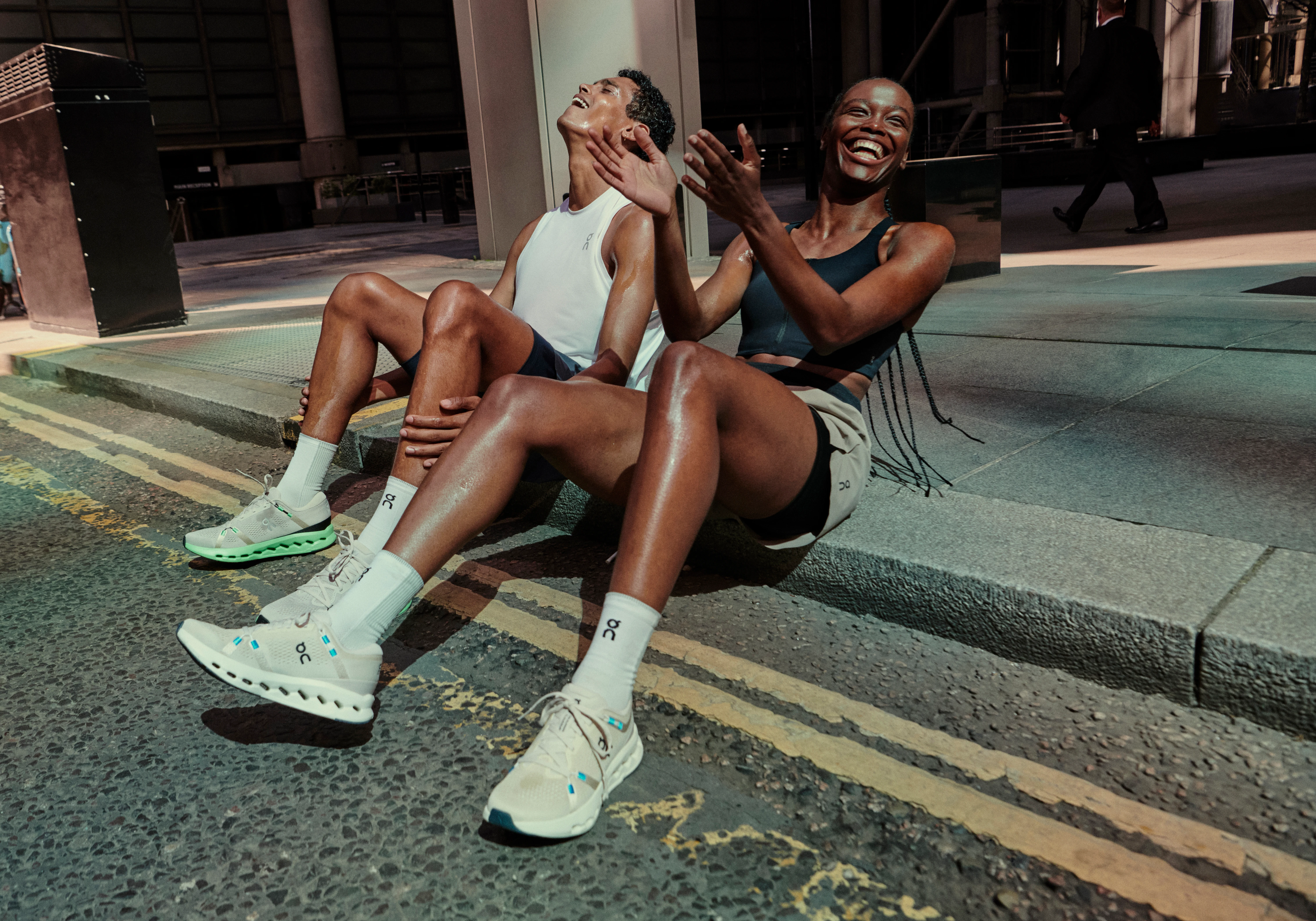 A man and a woman in On apparel sitting on the curb laughing.