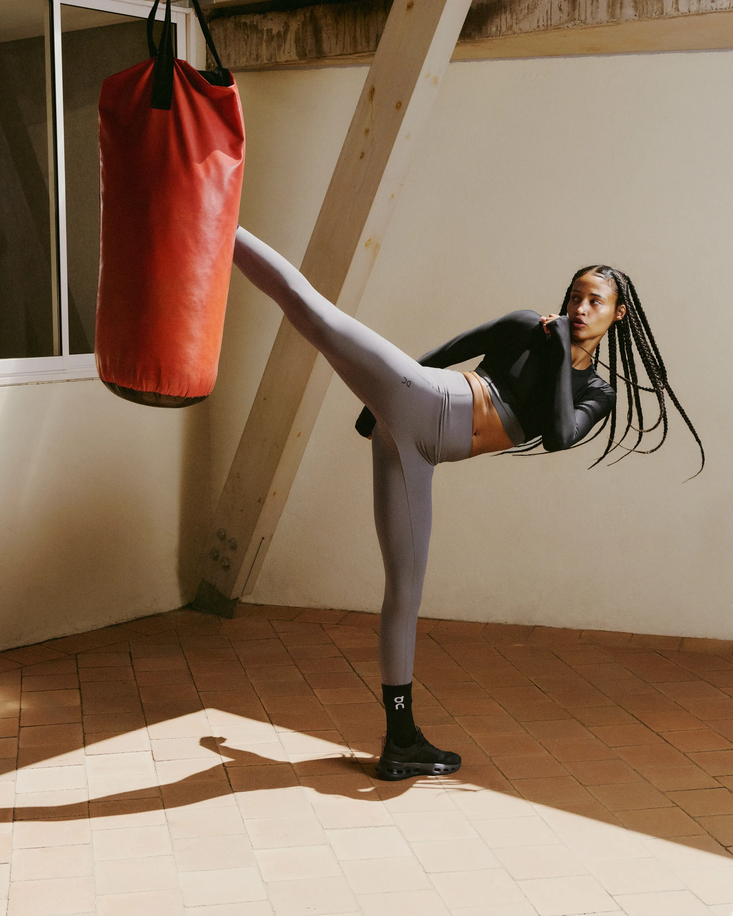 A female athlete practicing high kick on a punching bag while wearing On training apparel in a gym setting.