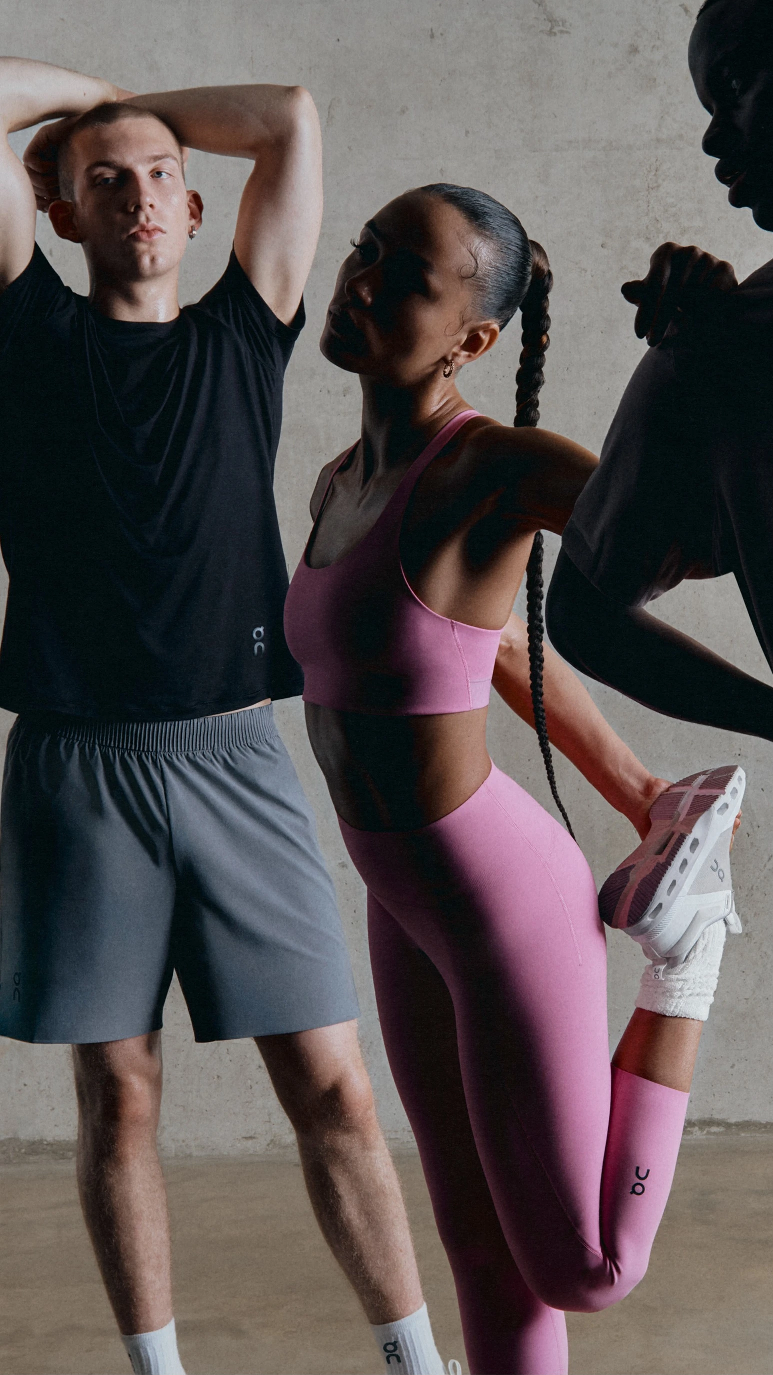 Three athletes in athletic wear stretch before performing physical activity. Two men are wearing neutral shorts and t-shirts, and the woman is wearing a pink sports bra and leggings.