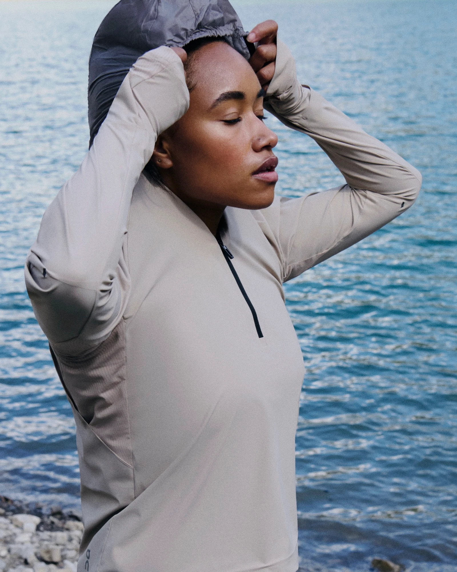 A woman with her eyes closed, standing beside a body of water, pulling her hood up.