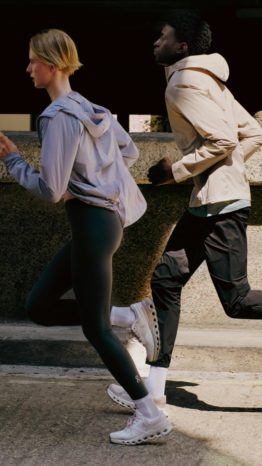 A woman runs slightly ahead of a man on a city sidewalk, both wearing On gear.