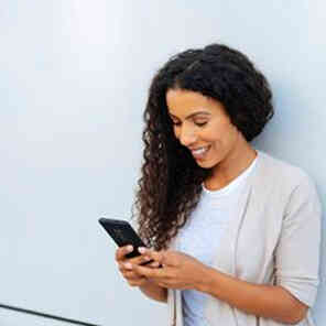 Woman smiling while using phone next to a wall.