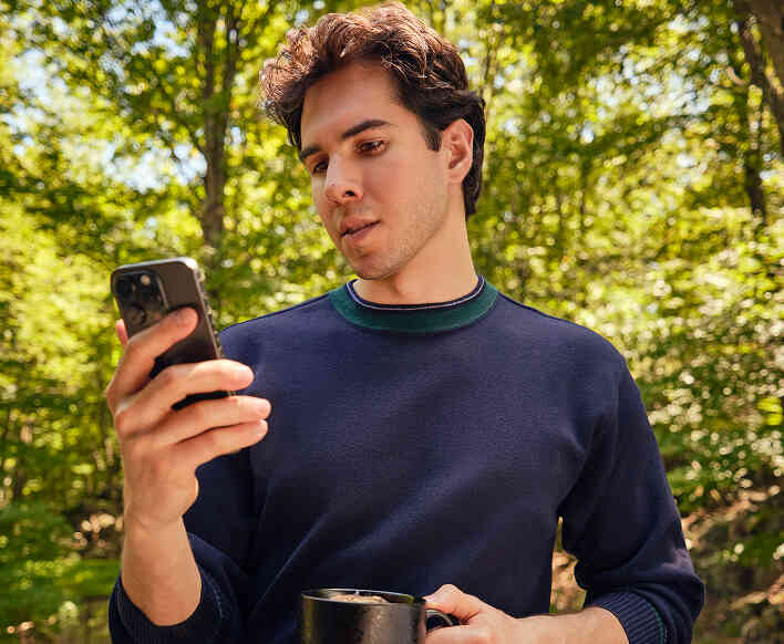 Man outdoors checking phone while holding a coffee.