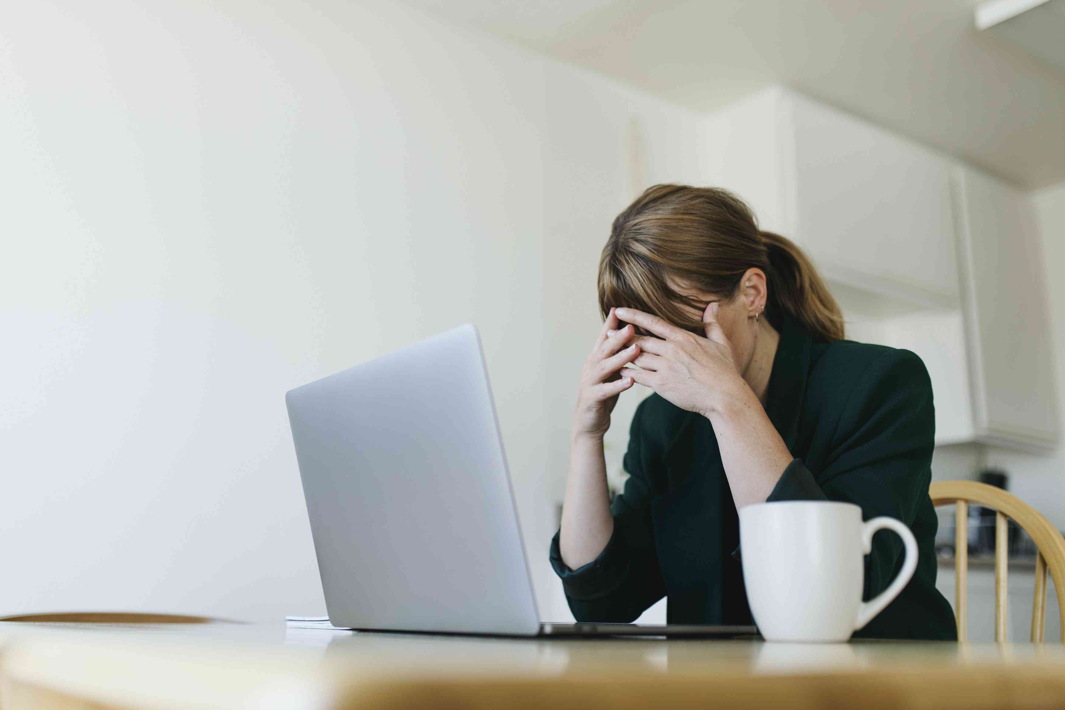 Woman covering face while sitting at laptop feeling overwhelmed.