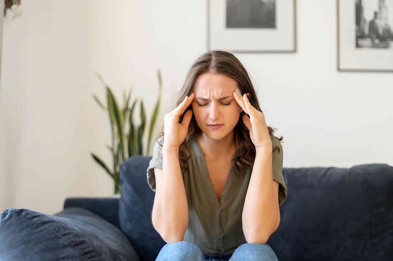 Woman sitting on couch holding temples with anxious expression.