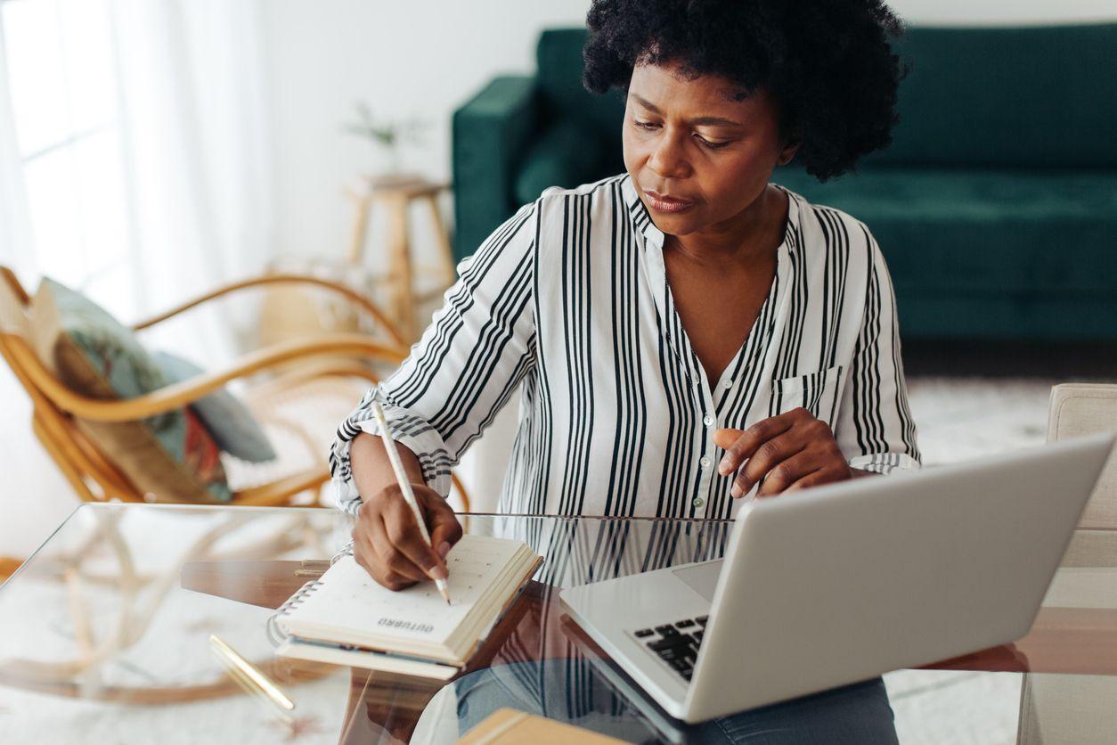 Woman writing notes while working on laptop at home.