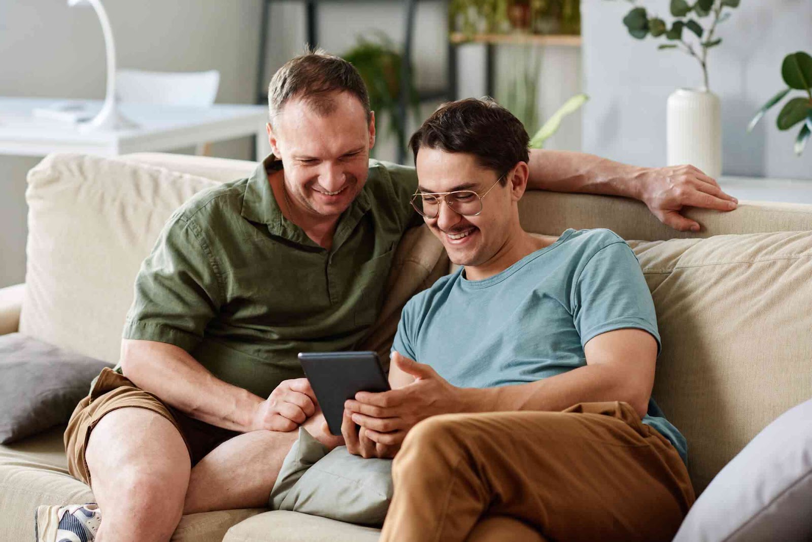 Two men smiling on couch using tablet together.