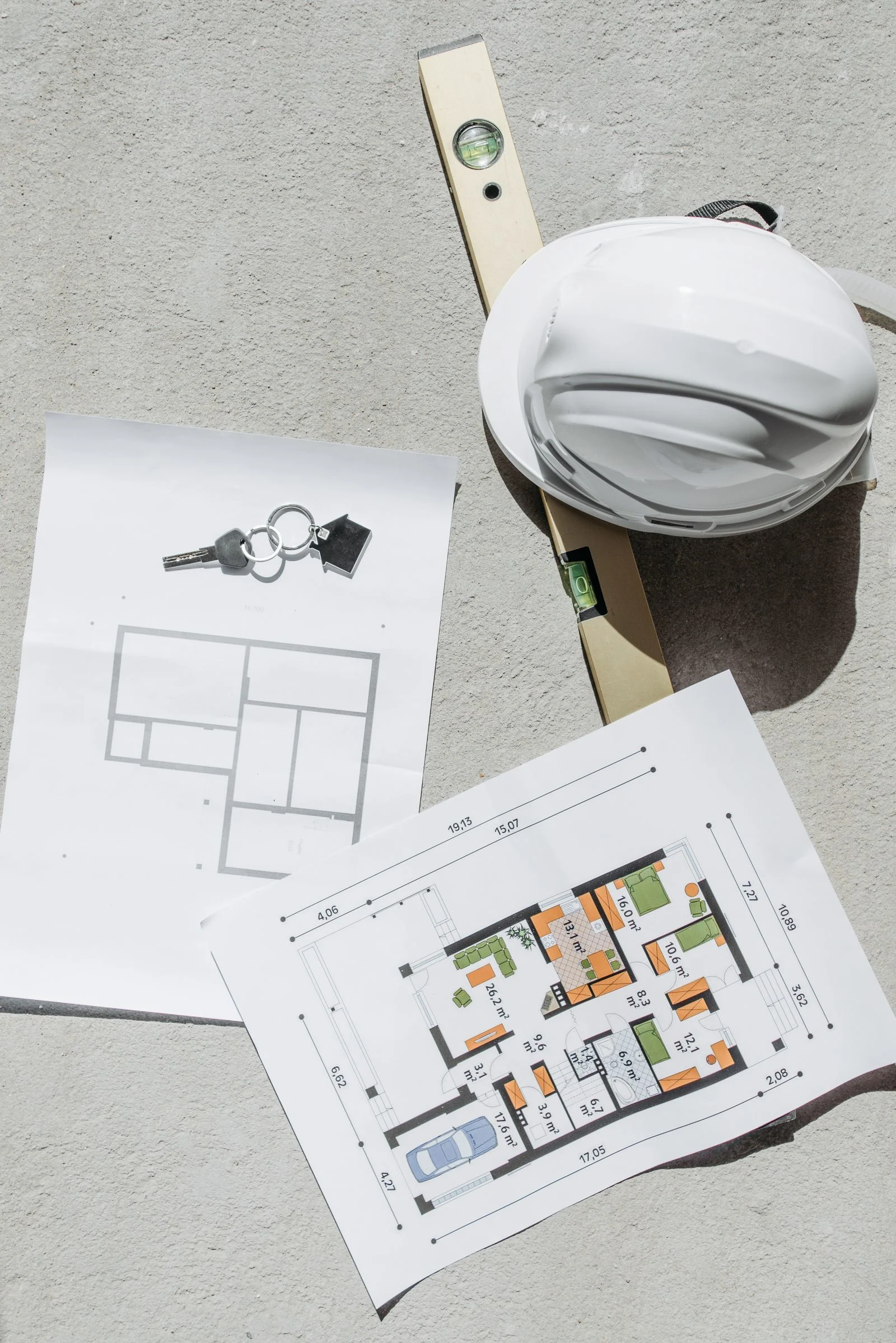 White construction hard hat, level, keys, and residential floor plans arranged on a neutral work surface
