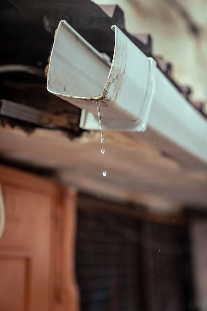 Water droplets falling from the open end of a metal gutter beneath a tiled roof
