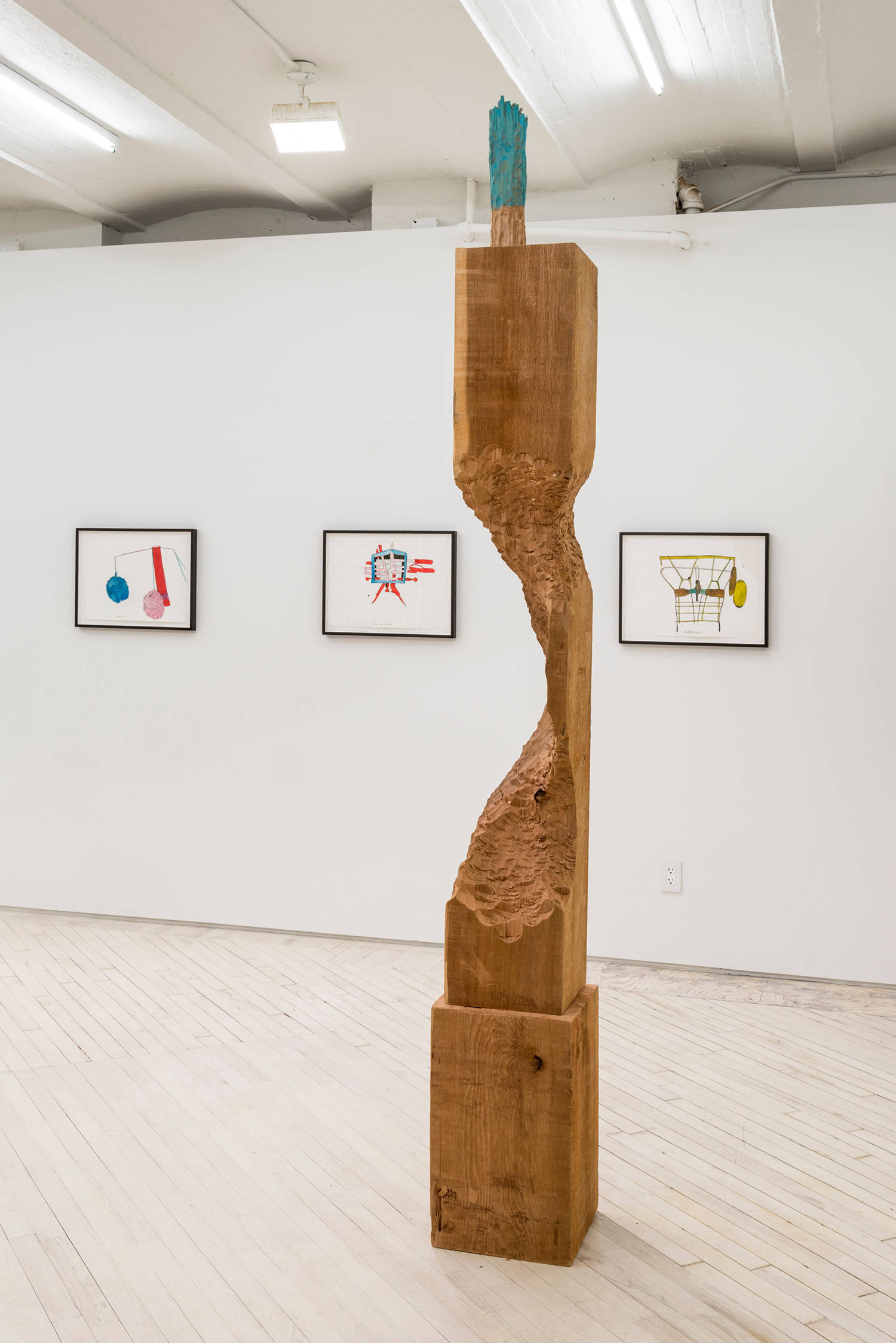 At the foreground a roughly carved tall wooden sculpture by David Adamo with a green painted object on the top, behind this are a suite of 3colorful ink drawings by Christine Rebet framed in black.