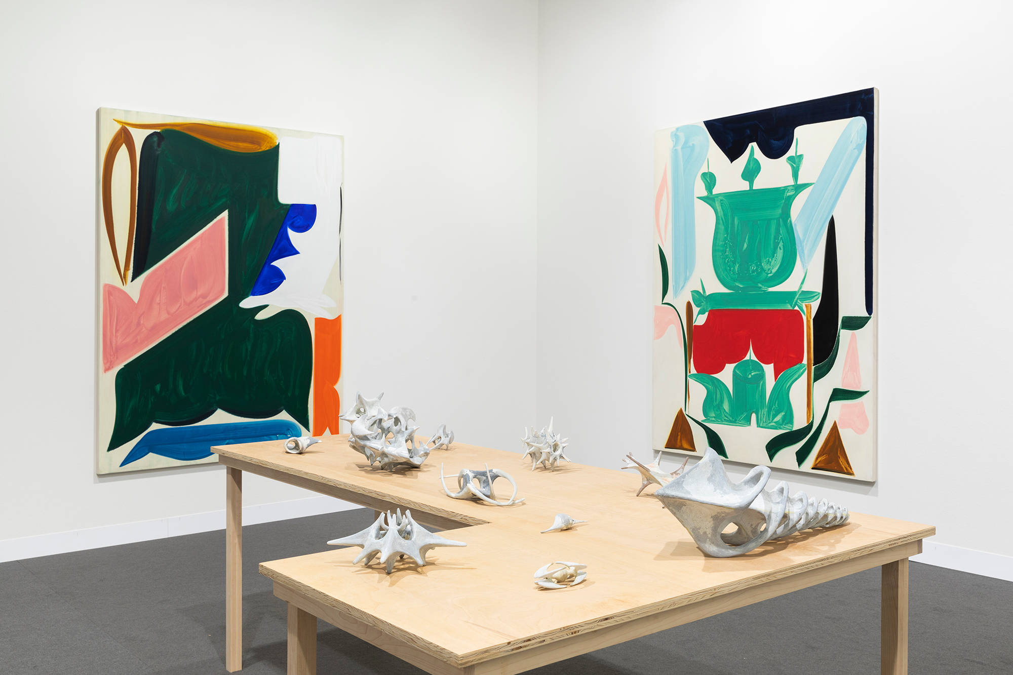In a gallery space, two large abstract paintings are spaced apart. The painting depict bold abstract geometric shapes in various hues of color. The dominant colors are hues of green, red, pink, blue, brown, white, and black. The paint is thin and gestural. In the center of the gallery space is a freestanding brownish table with various miniature gray sculptures placed evenly apart. The sculptures are abstract and resemble organic material or bone formations.
