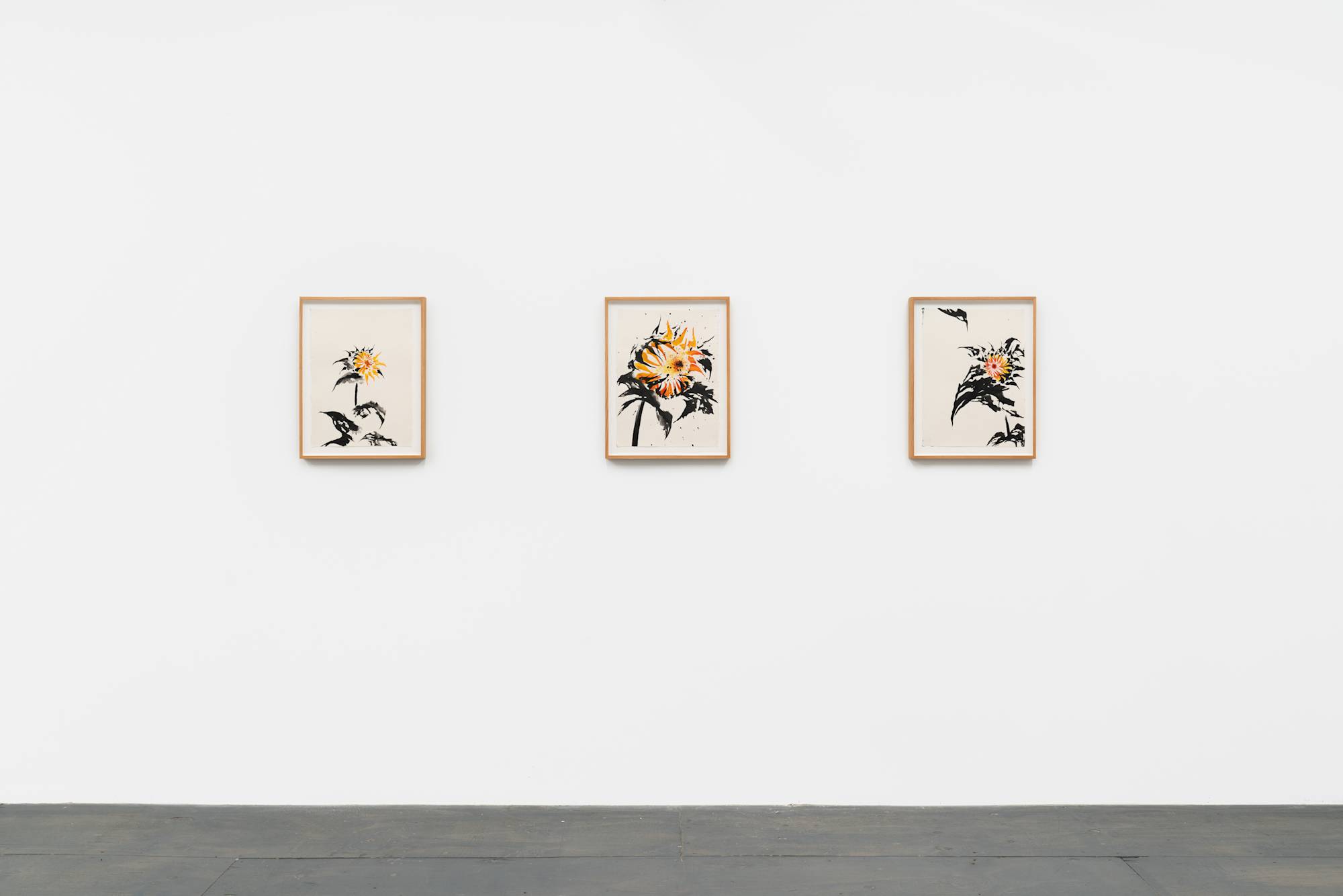 Three small mounted ink paintings of sunflowers with yellow-orange petals and black leaves and stems hang on a white wall.