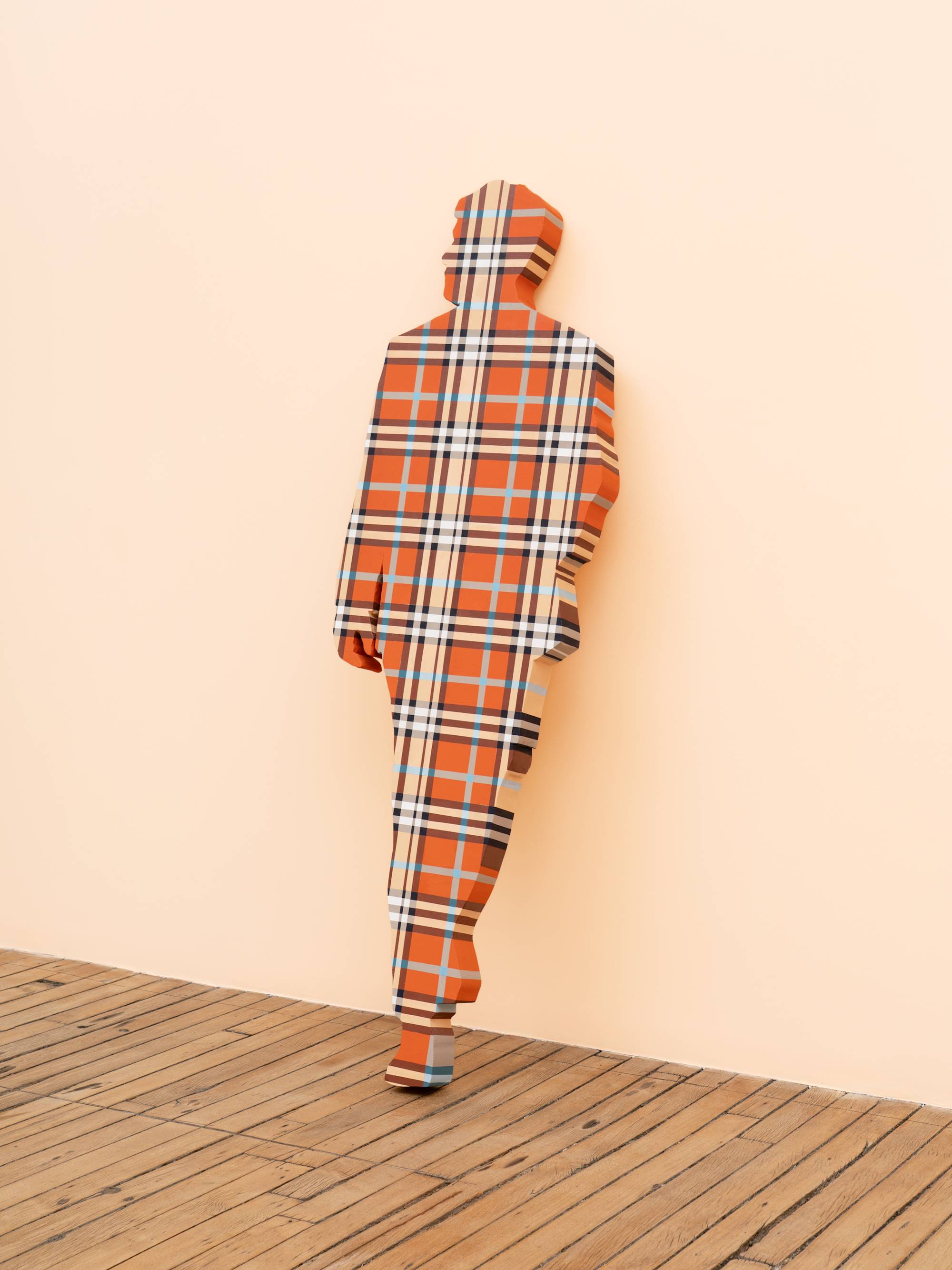 A life-sized human figure painted in a blue and orange plaid pattern leans against a peach wall in an art gallery.