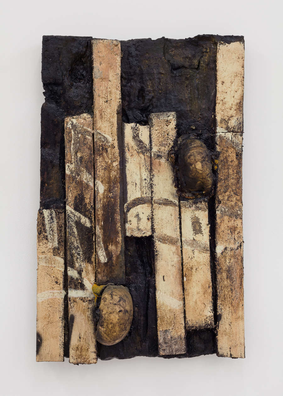 Image of a sculpture by Brandon Ndife. Hanging on the wall of a gallery is what appears to be a panel with wooden planks (like a wooden floor was excavated and hung upright on the wall). The wood planks are very dirty with graffiti marks on them and round gourd like object growing on the surface.