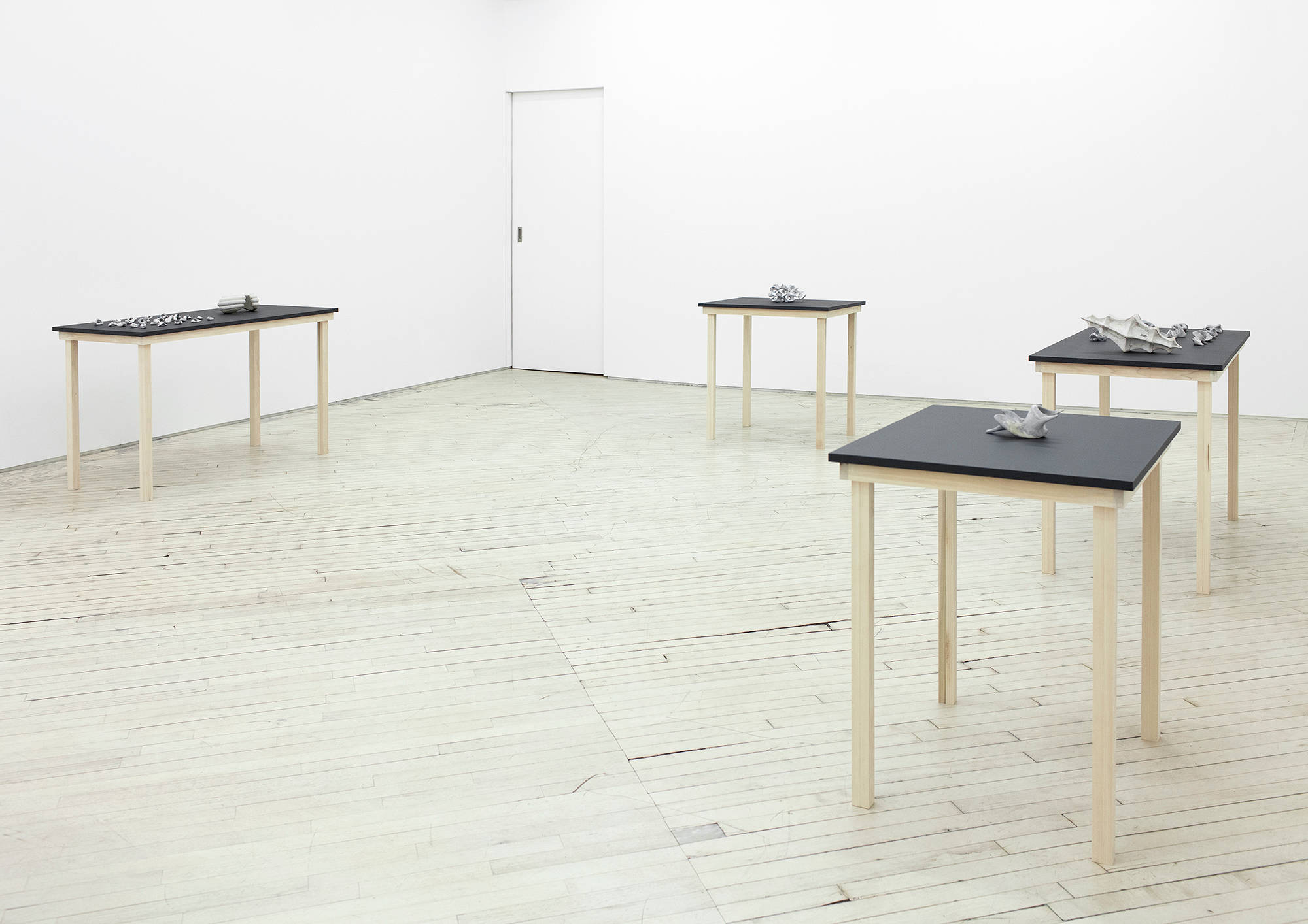 Installation view of several sculpture in a gallery by Matt Hoyt depicting black table with wooden legs upon which hand made sculptures sit. There are tables with many sculptures and tables with only one sculpture.