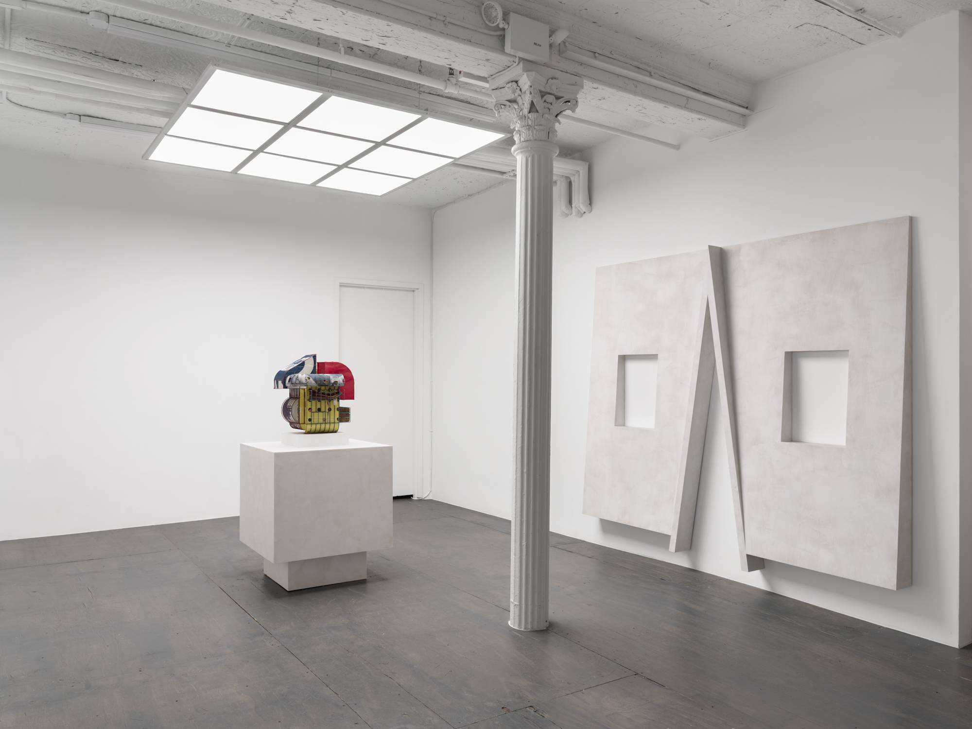 Angle view of a white walled gallery with grey floor and white column lit by an LED light fixture that resembles a skylight. Two sculptures are visible in the gallery. One large wall mounted sculpture on the wall to the right and one standing on a pedestal in the center of the room.