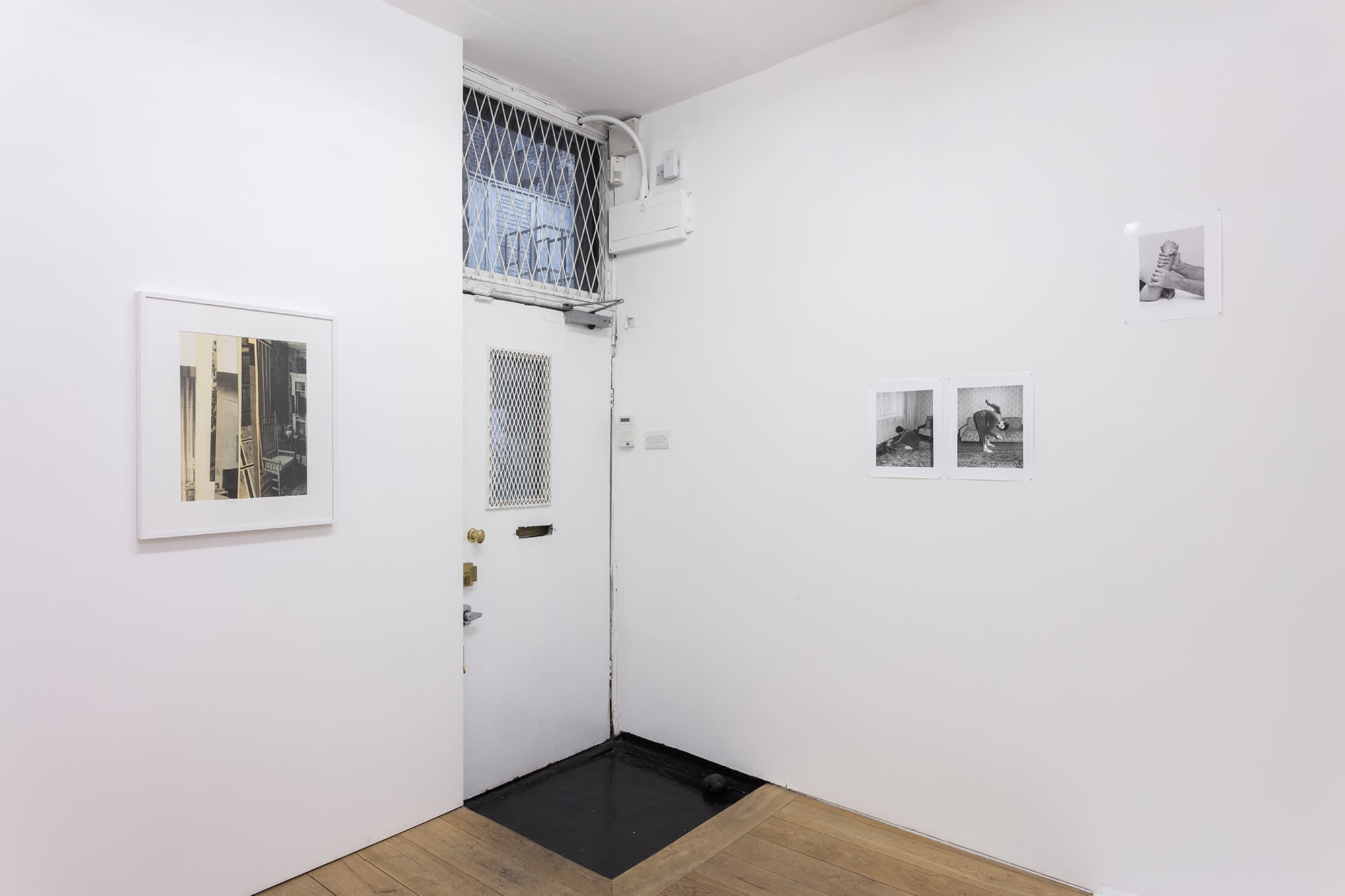 Installation view of a front door painted white and photographs installed to the right and left of the door. To the left is an abstract color photograph, framed and matted in white, with predominantly tan, yellow and black colors. The image has many vertical lines and on the right hand side we seen an interior with chair. To the left are three black and white photos, two in a pair, showing people in highly textured interior spaces in strange poses and one above to the right shows two hands of a white person on gripping the white forearm of another person.