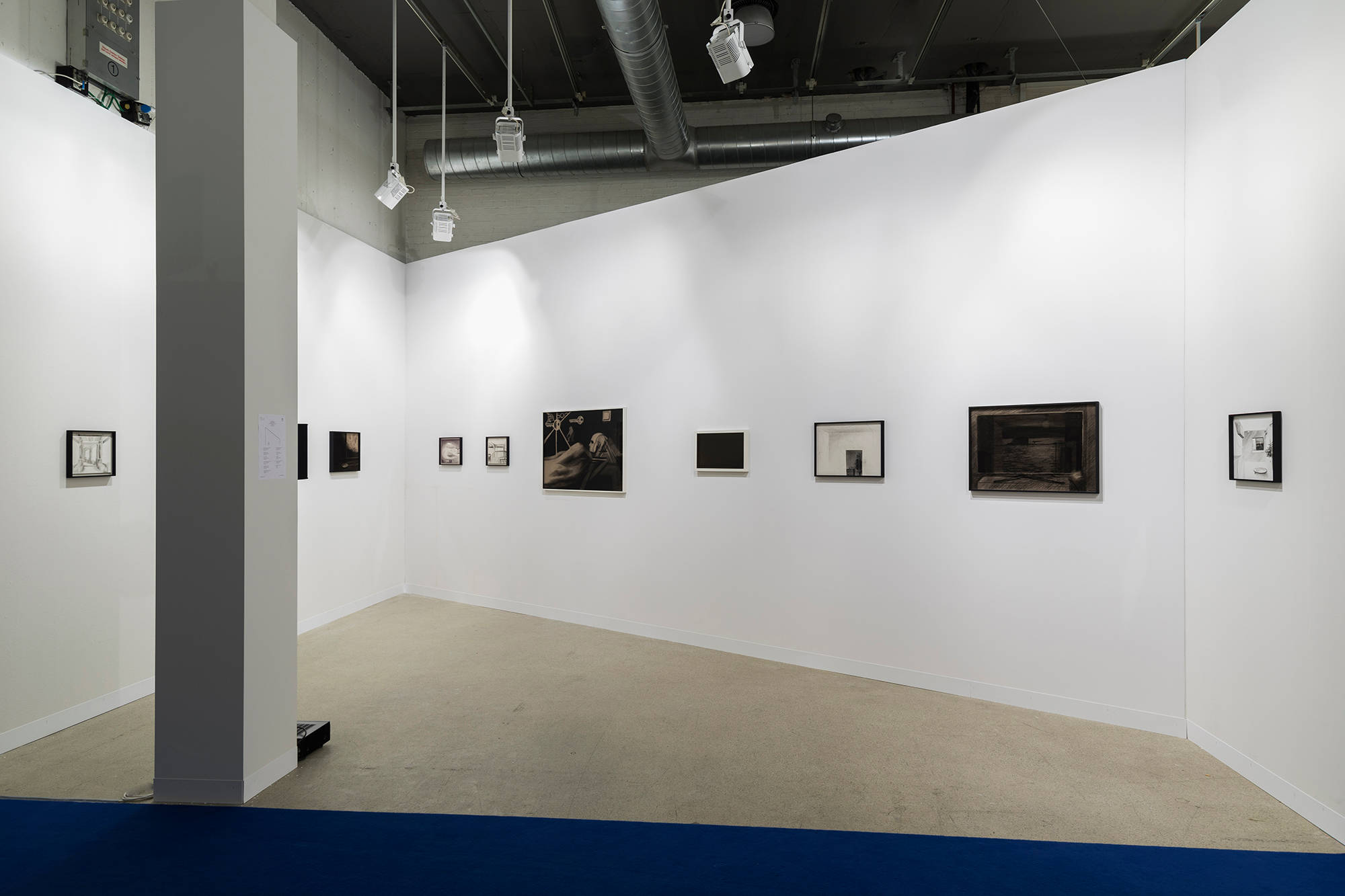 Installation view of an art fair booth with an angled back wall, framed drawings hung in a row on all three walls of the booth.