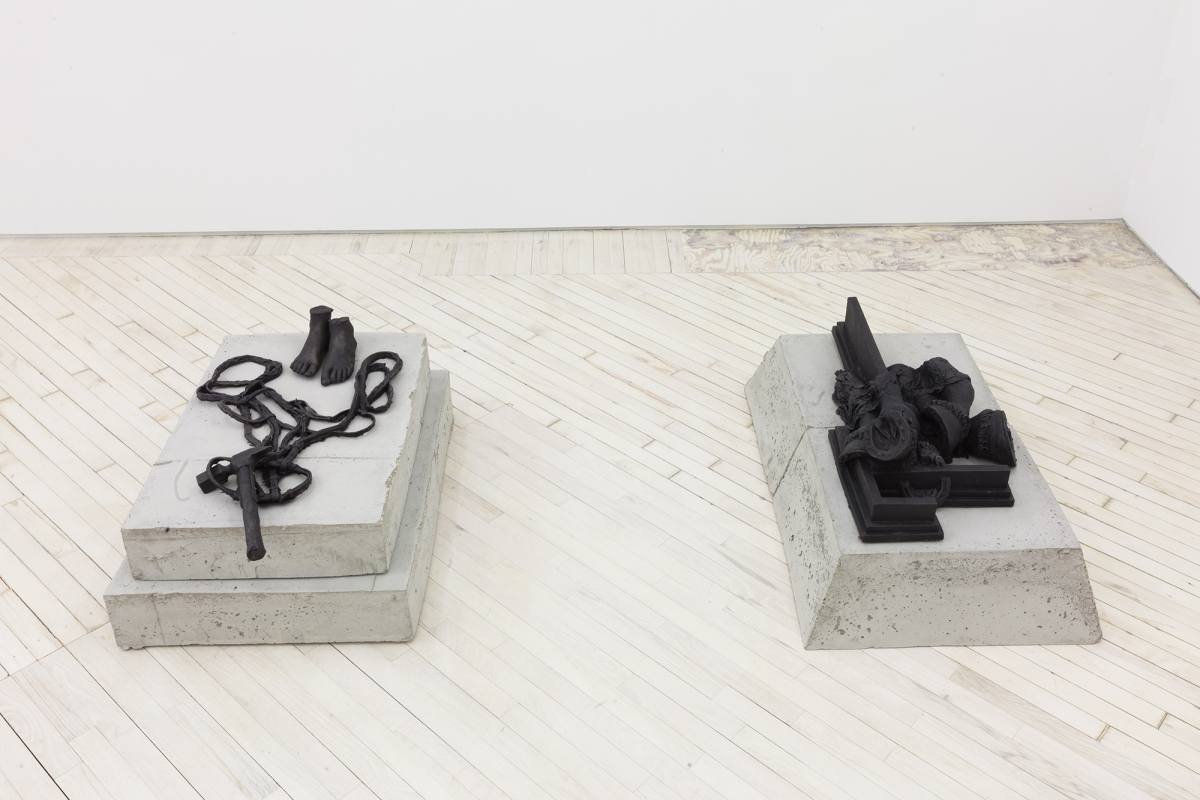 In a gallery space, two cast iron sculptures depicting a rope, human feet, and an architectural structure resting on top of concrete plinths. 