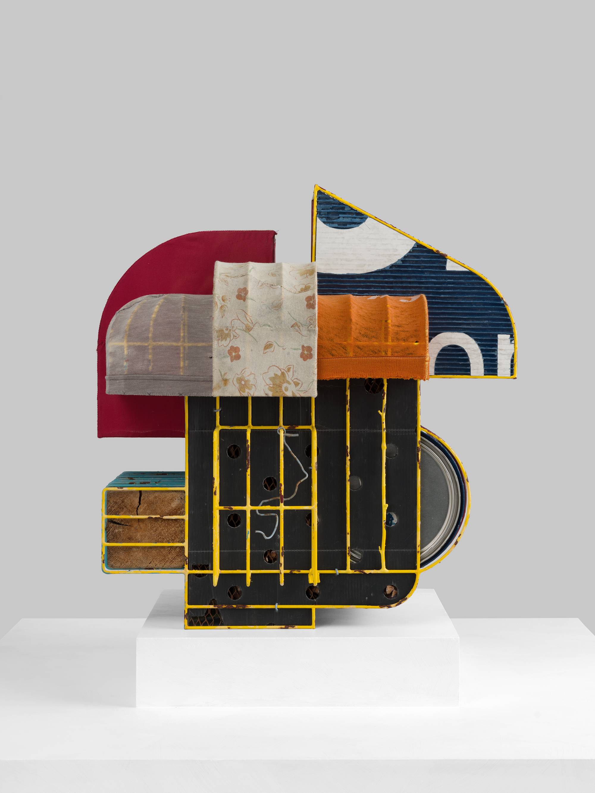 A cage-like colorful steal wire sculpture, wrapped with several tattered fabrics to resemble various awnings, and embellished with an old shoe, a twig, printed coroplast signage, an old blue paint can, a lock, a paper tag, and a wooden block sits on a white pedestal against a white wall.