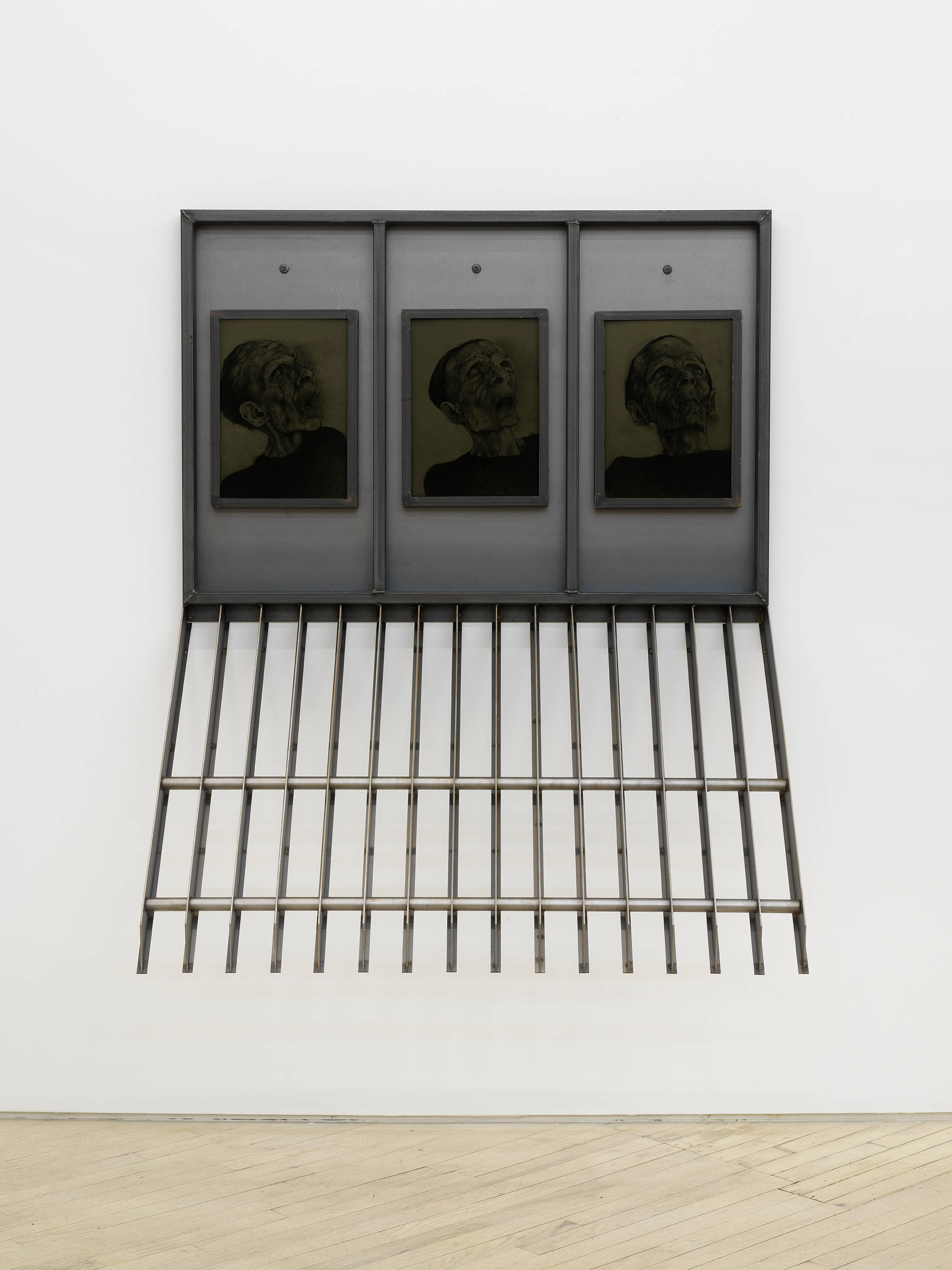 A large steel sculpture hanging from a wall containing three drawings a human head.