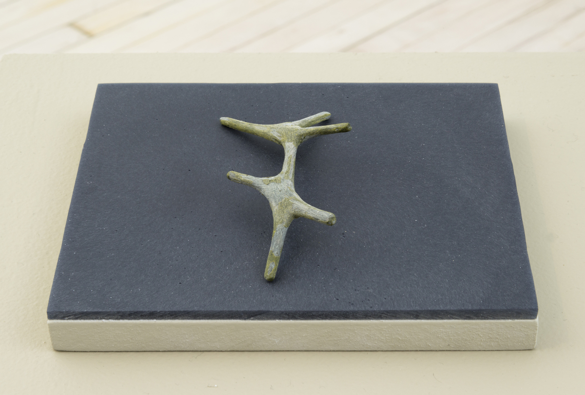 An abstract sculpture resembling a bone or other organic material resting on top of a dark gray plinth. A light brown floor is partially visible in the background. 