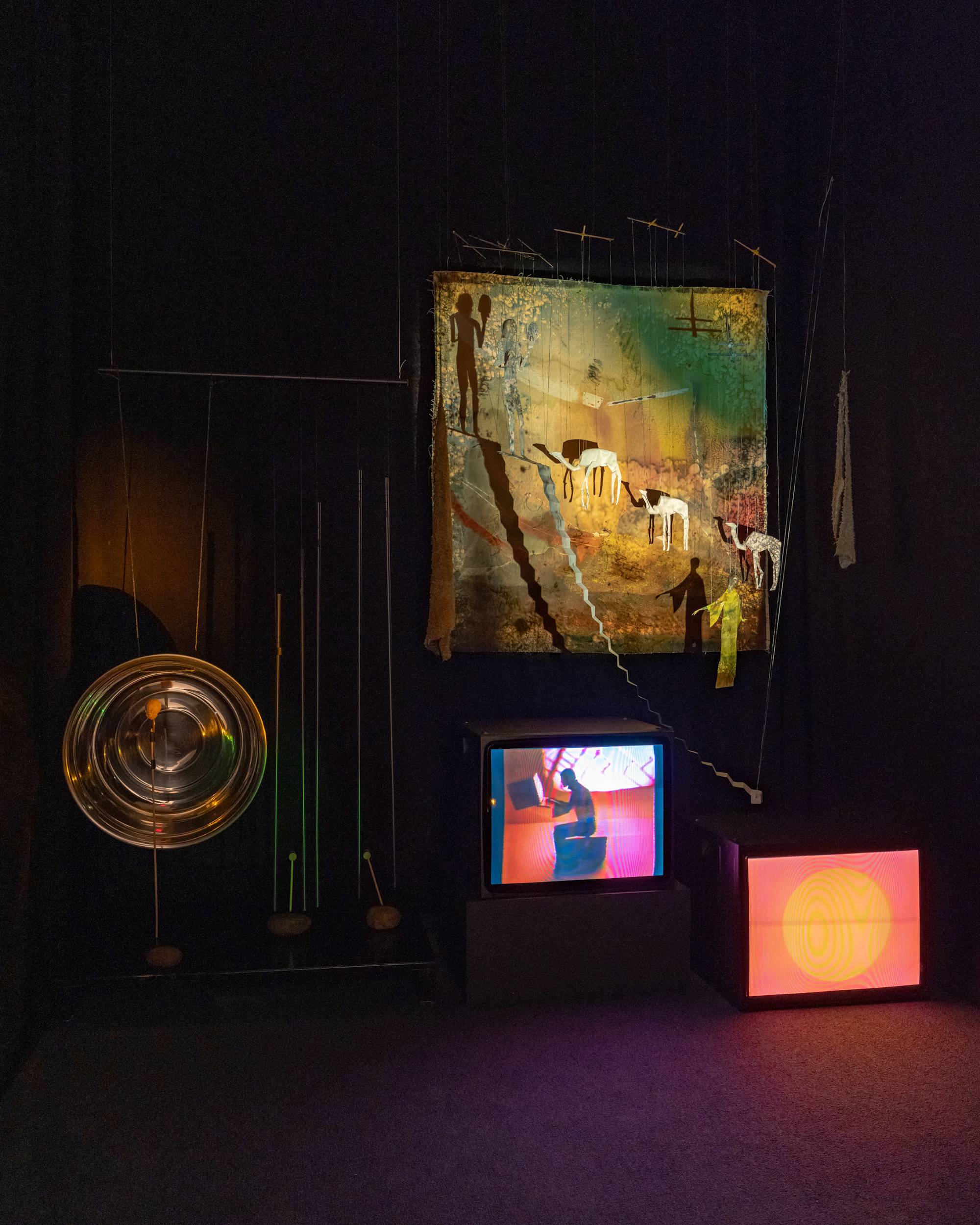 A dark room illuminated by a single projector and the glow from two CRT monitors. The monitors, set close to the floor, feature abstract animated videos. Above the monitor string puppets figures of 2 humans, 3 camels and a staircase are suspended in front of an abstract painting. To the left, a make-shift gong and wind chimes are suspended from the ceiling.