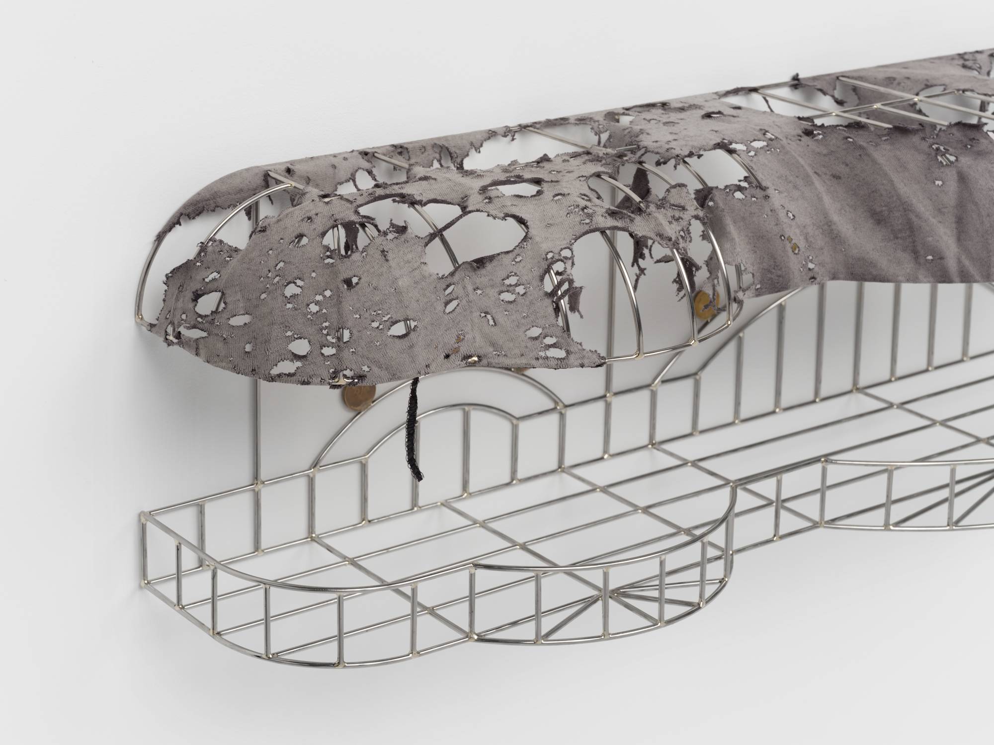 Detail of a cage-like chrome sculpture, wrapped with a tattered grey t-shirt to resemble an awning, and embellished with soldered on gold-colored coins.