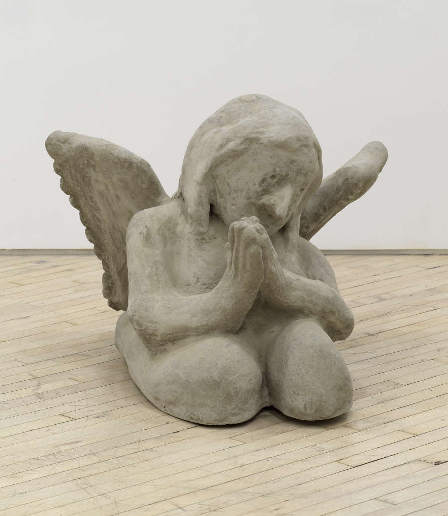 Image of an artwork by Libby Rothfeld of a cartoonishly rendered angel seated on the floor with wings and hands in a prayer like pose. The angel is sculpted from concrete.