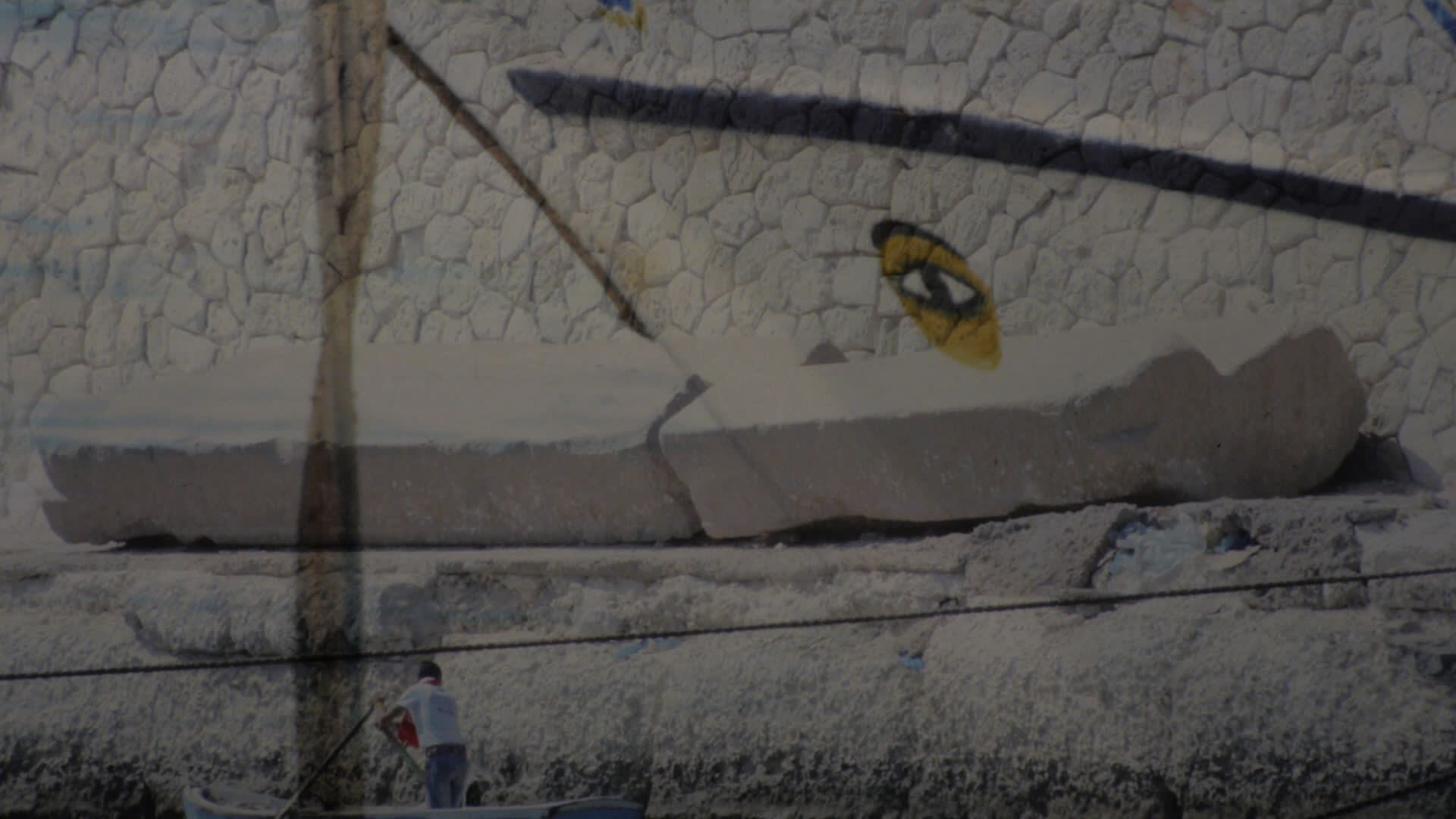 Image from Ellie Ga's video 'Four Thousand Blocks' showing stone ruins on a harbor in Alexandria of pale stone and concrete, a painted eye with a yellow circle around it is at the center/right.