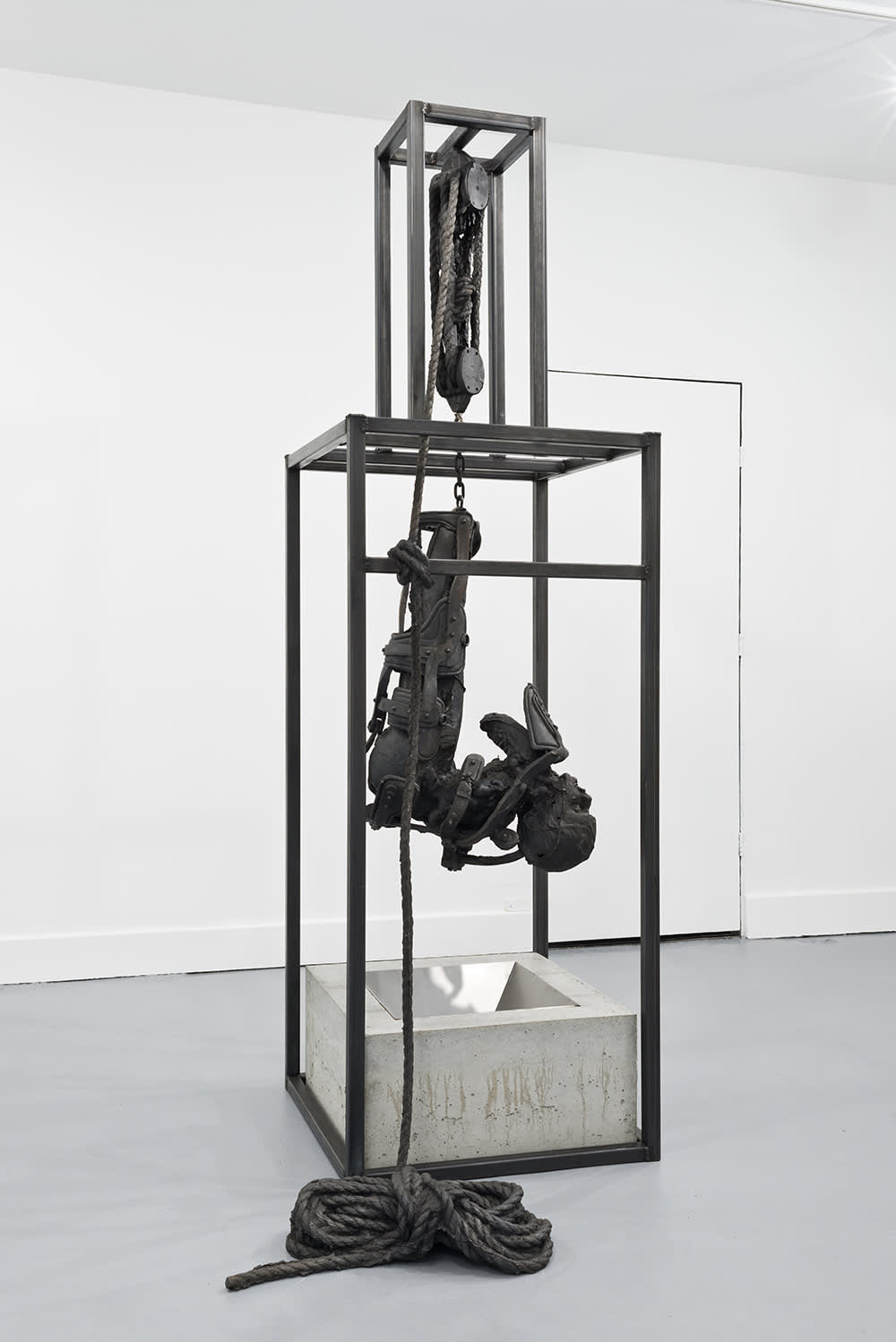 Installation view of a metal sculpture of a vertical tower, with a sculptures made of cast iron depicting a a figure in agony suspended inside of the tower by with cast iron ropes. There is a concrete and metal trapezoid at the base of the sculpture. 