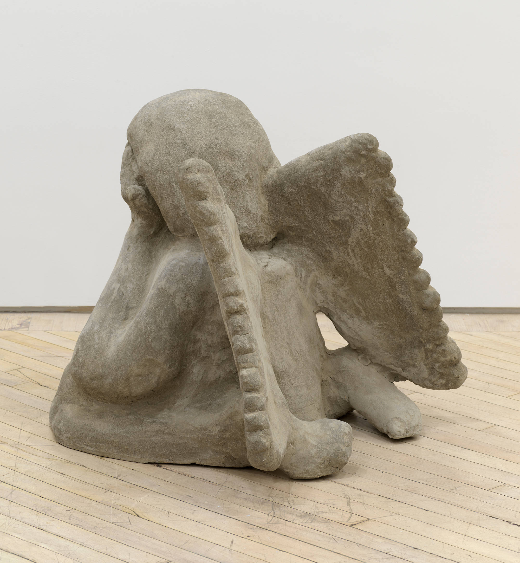 Image of an artwork by Libby Rothfeld of a cartoonishly rendered angel seated on the floor with wings and its hands covering its eyes. The angel is sculpted from concrete.