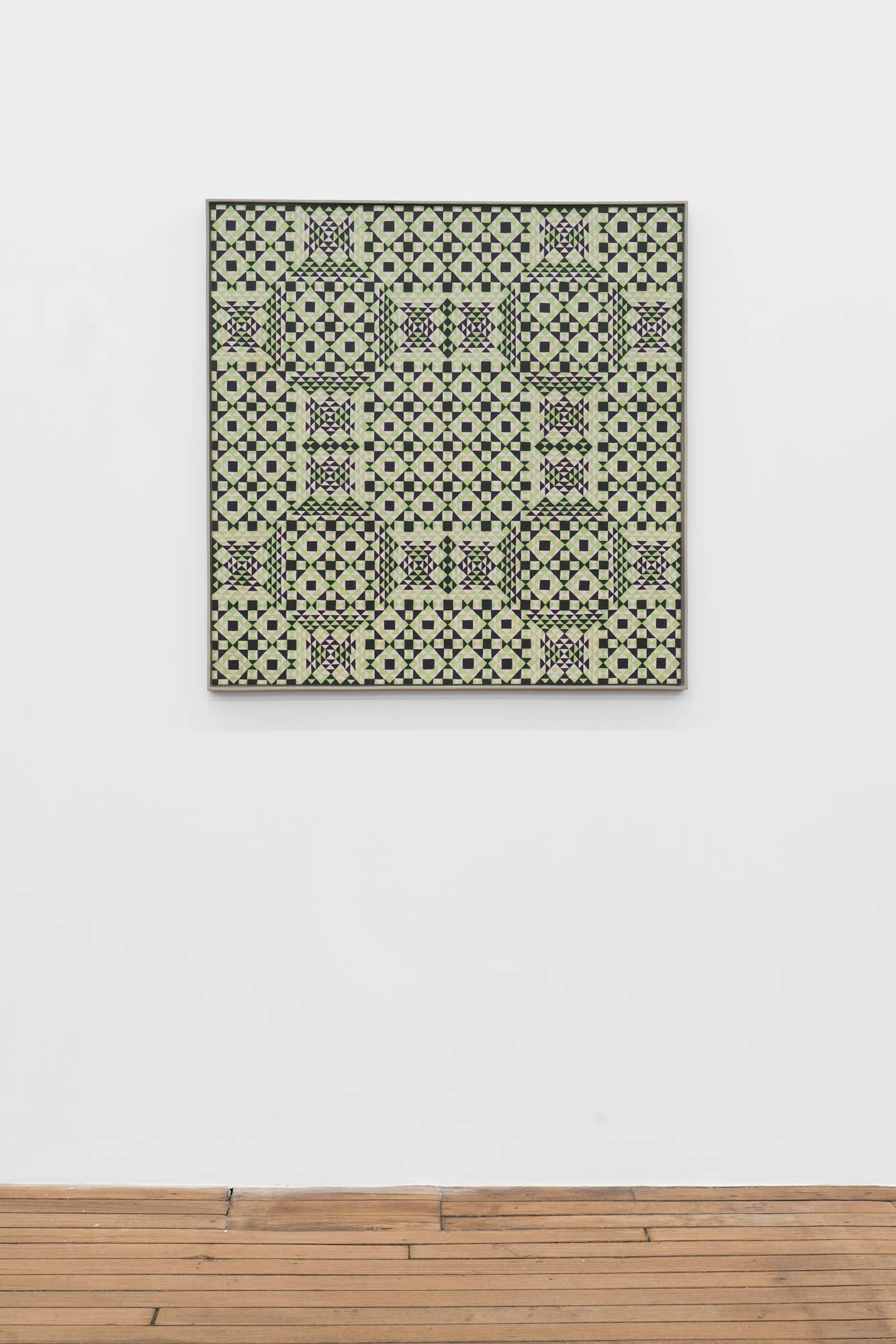 A colorful, symmetrical, geometric painting on board consisting of a pattern of repeating diamonds and triangles of green, light green, and dark green, in a light green artist frame hangs on a white wall over a wood floor.