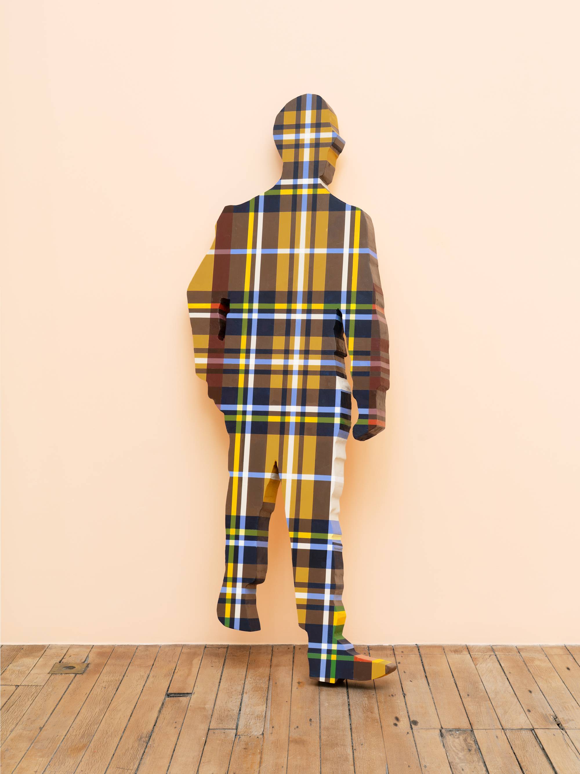 A life-sized human figure painted in a brown, yellow and blue plaid pattern leans against a peach wall in an art gallery.
