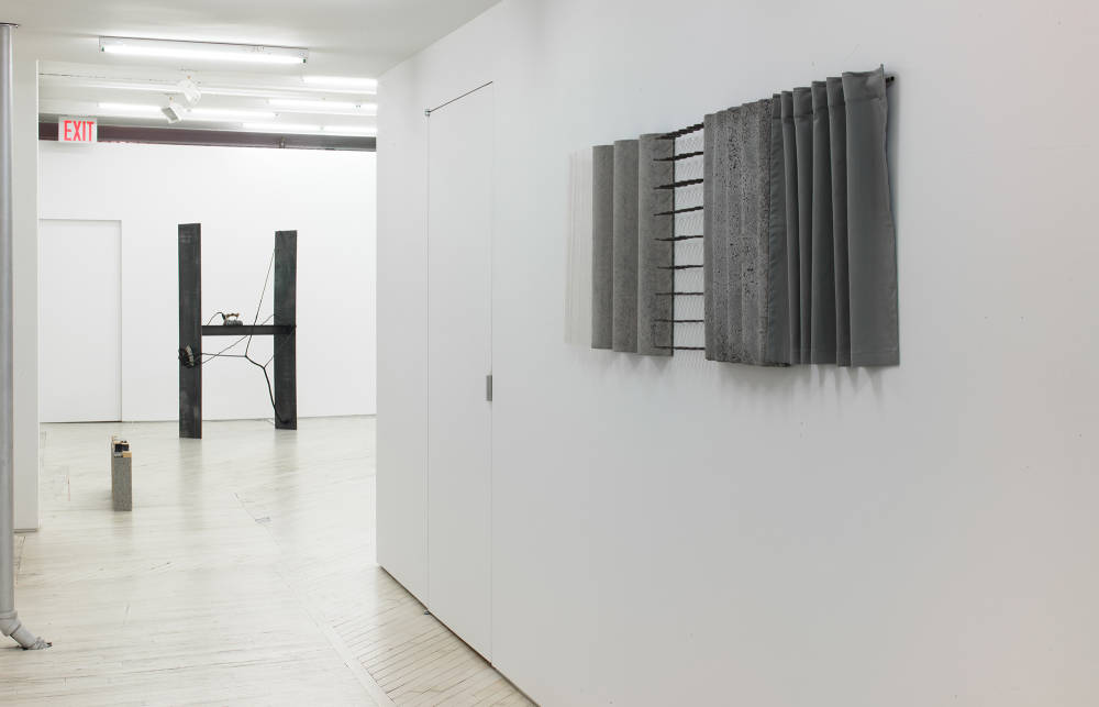 In the hallway of a gallery space, a horizontal wall sculpture is hung on the right wall. The sculpture contains rows of industrial materials including concrete and steel. In the background is a larger gallery space containing a large metal sculpture of irregular shape.