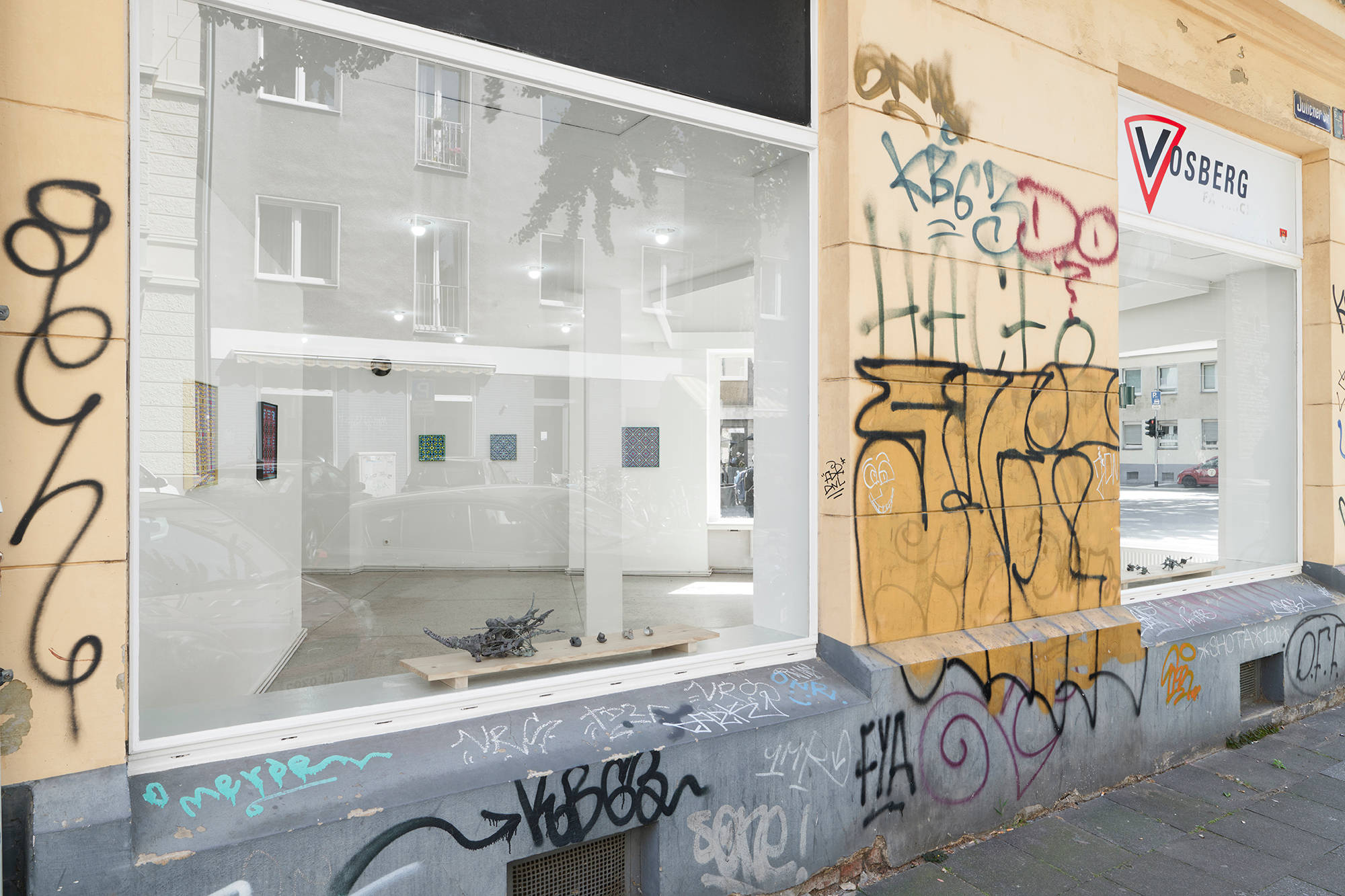 Outside view of the gallery "Vosberg" with a yellow wall that has graffiti covering it. Through two large windows, you can see the exhibition from outside. You can see five paintings.