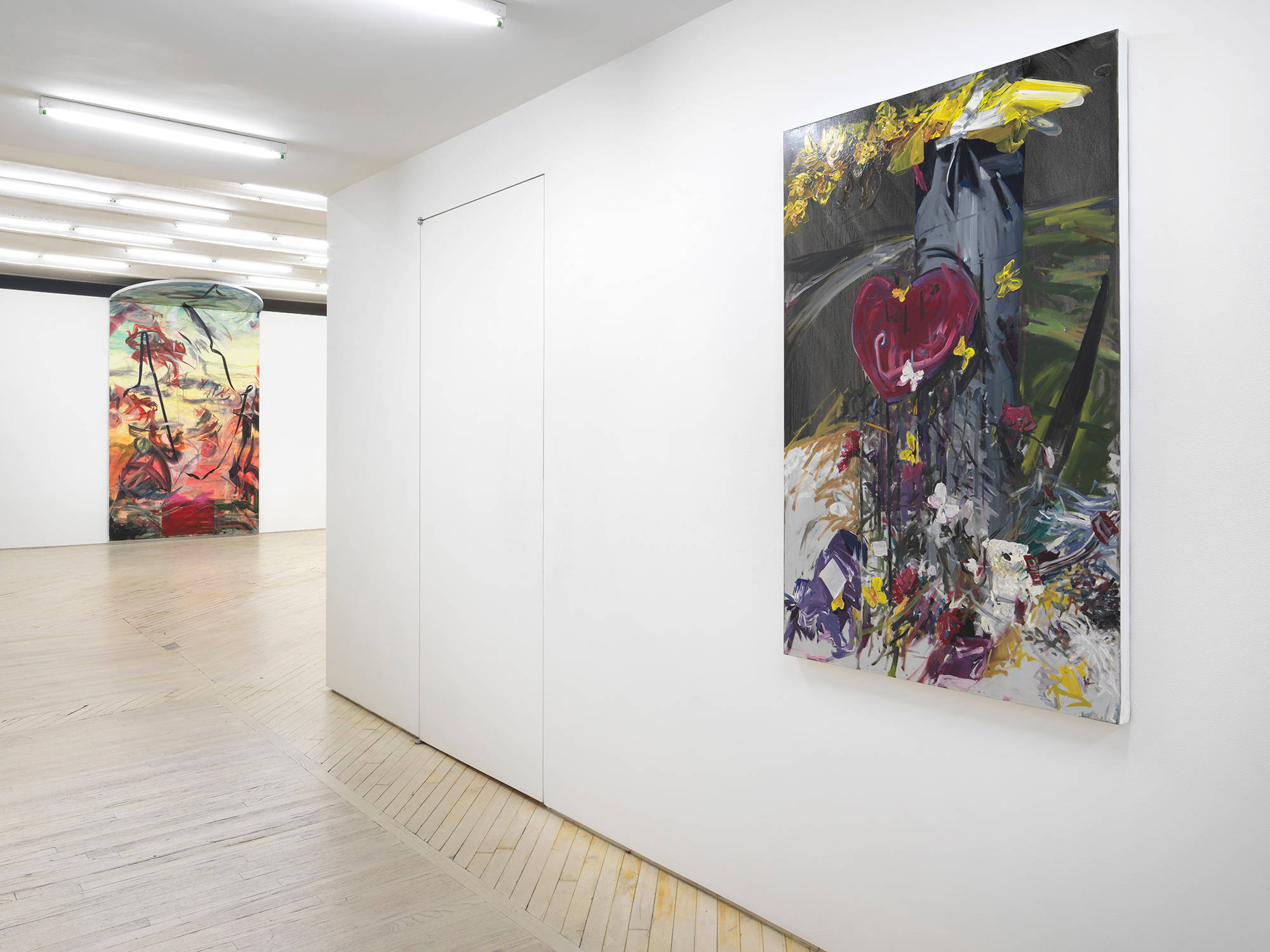 In a gallery hangs two paintings by Tom Holmes, one on a hallway wall that depicts a scene of a roadside memorial with flowers resting on the sidewalk next to a street lamp, and another large columned painting that touches the ceiling with mostly abstract painterly marks.